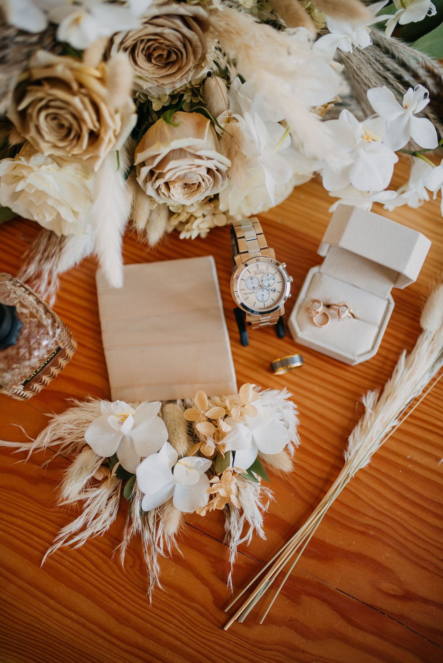 Flat lay of wedding details including roses, watch, ring, and floral boutonniere on wood surface.