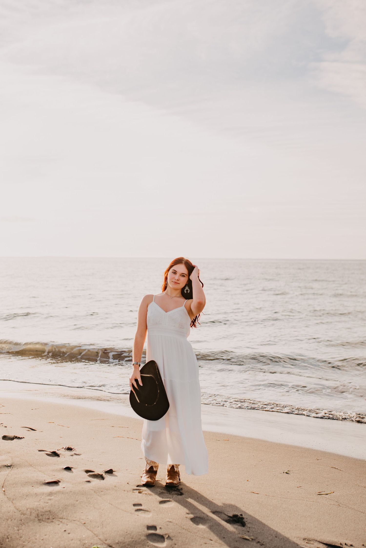 Elegant woman in white maxi dress holds black hat while standing on a sandy beach.