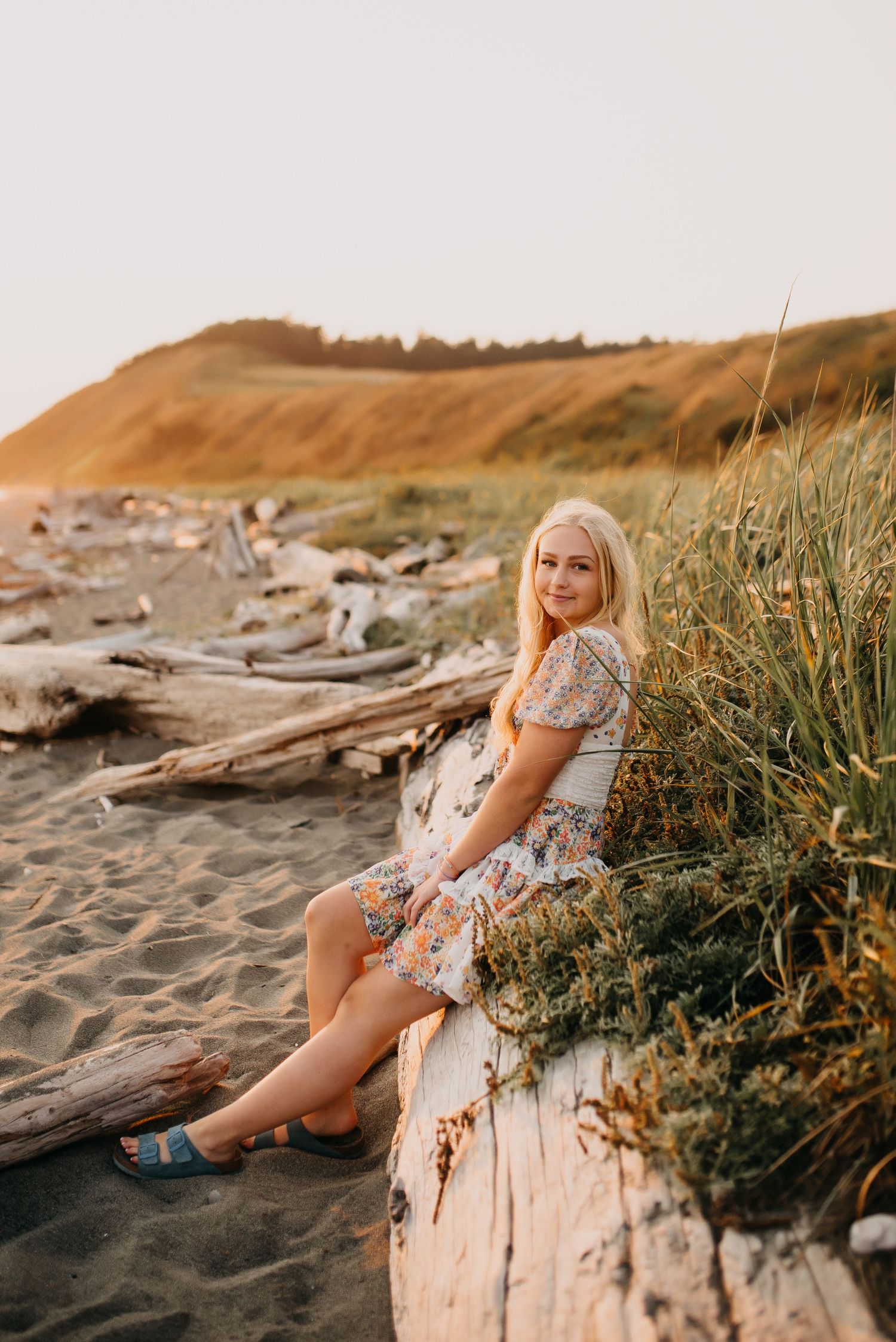 Blonde woman in floral dress sits on driftwood near beach grass with sandy dunes glowing in sunset light.