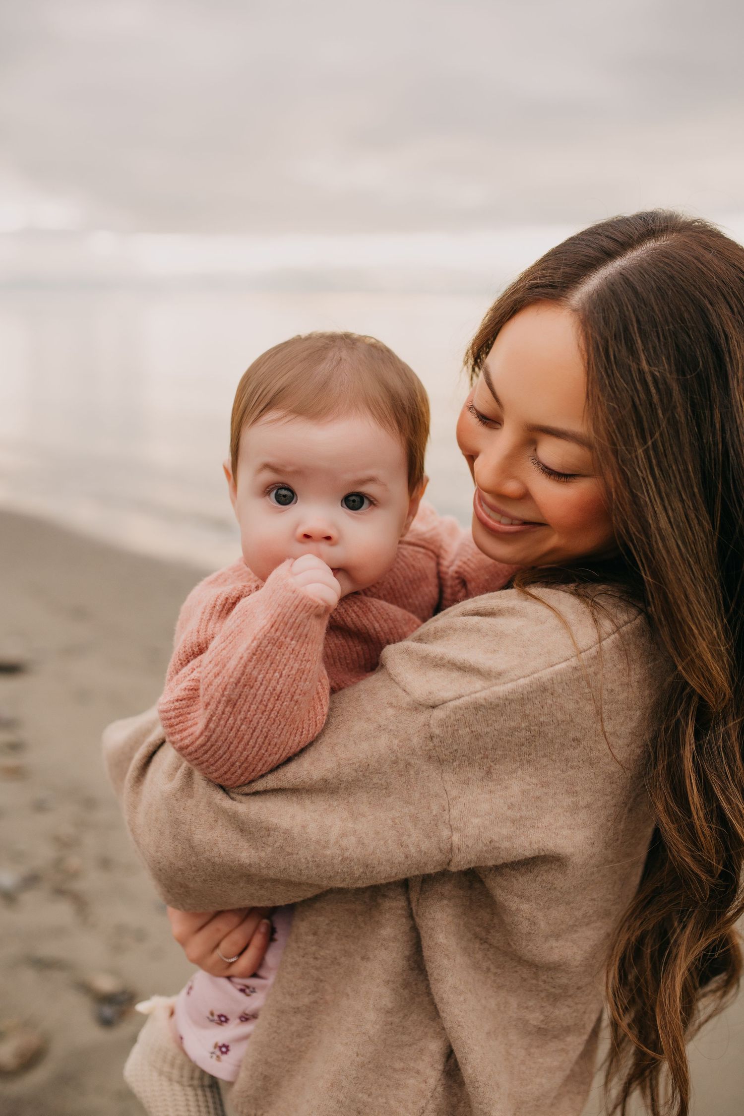 Smiling brunette mother holding her baby girl in a pink sweater on a beach.