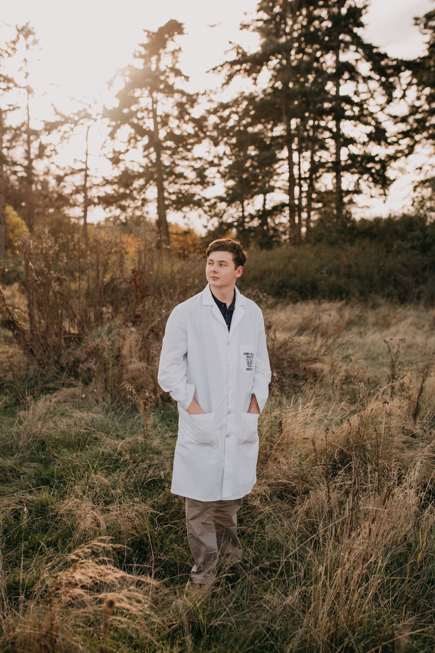 Young man in white lab coat standing in autumn field with pine trees for senior portrait.