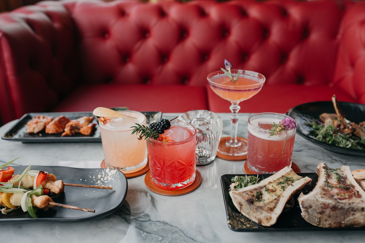 Colorful cocktails and appetizers displayed on a marble table with red tufted booth.