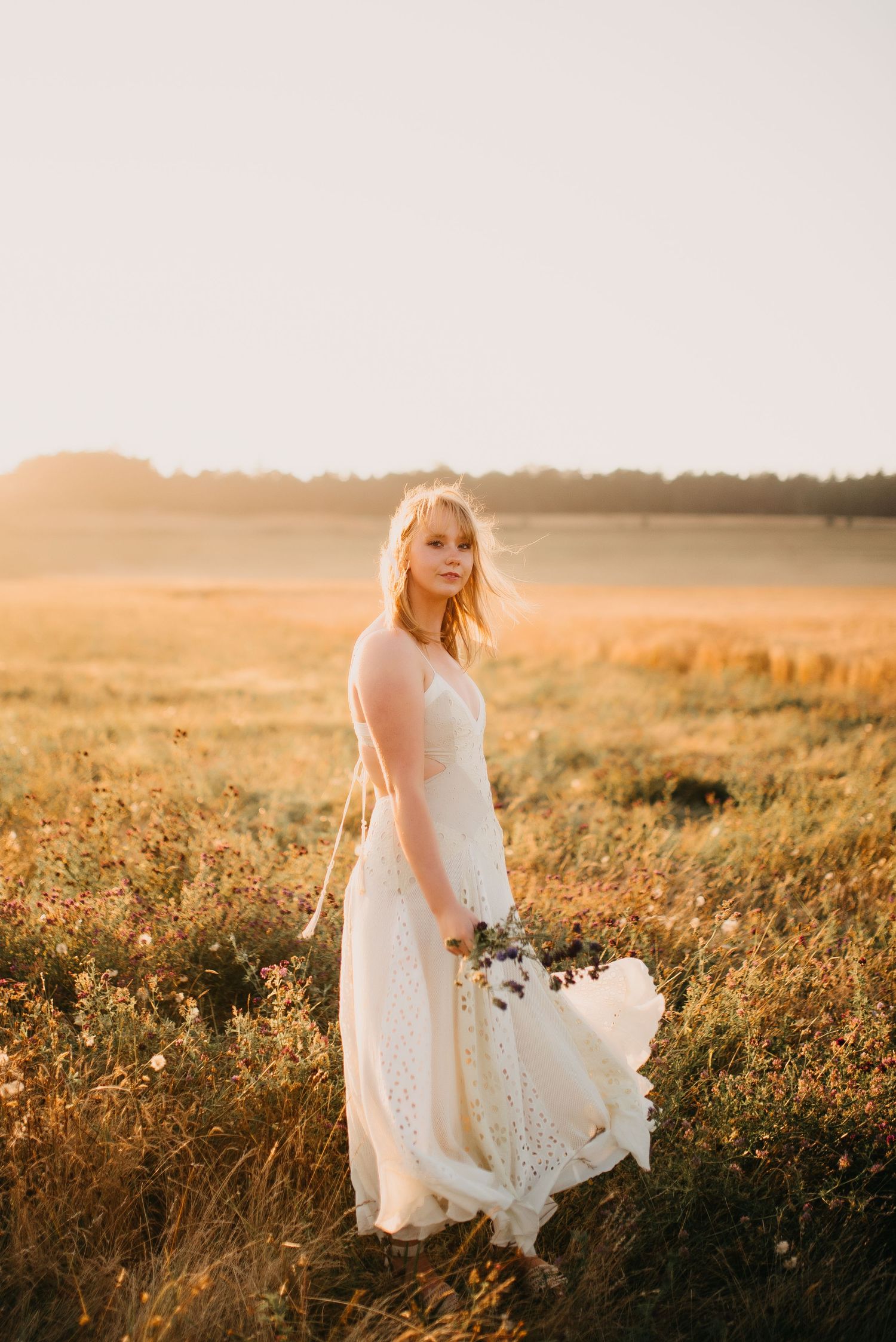 Blonde woman in white lace bridal gown holding wildflowers in a sunlit golden field at sunset.