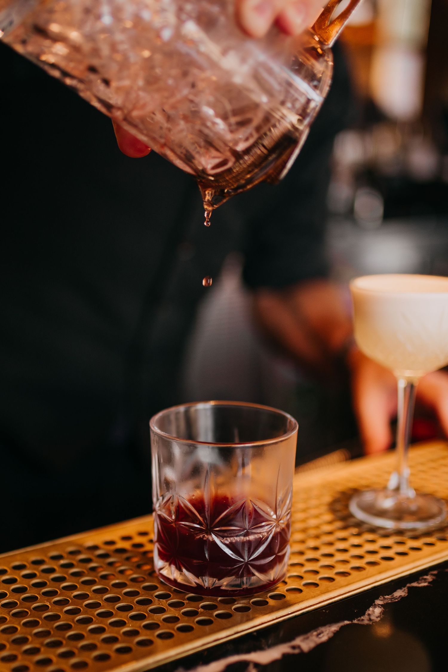 Bartender pouring dark cocktail from crystal pitcher into a rocks glass on a gold bar mat.