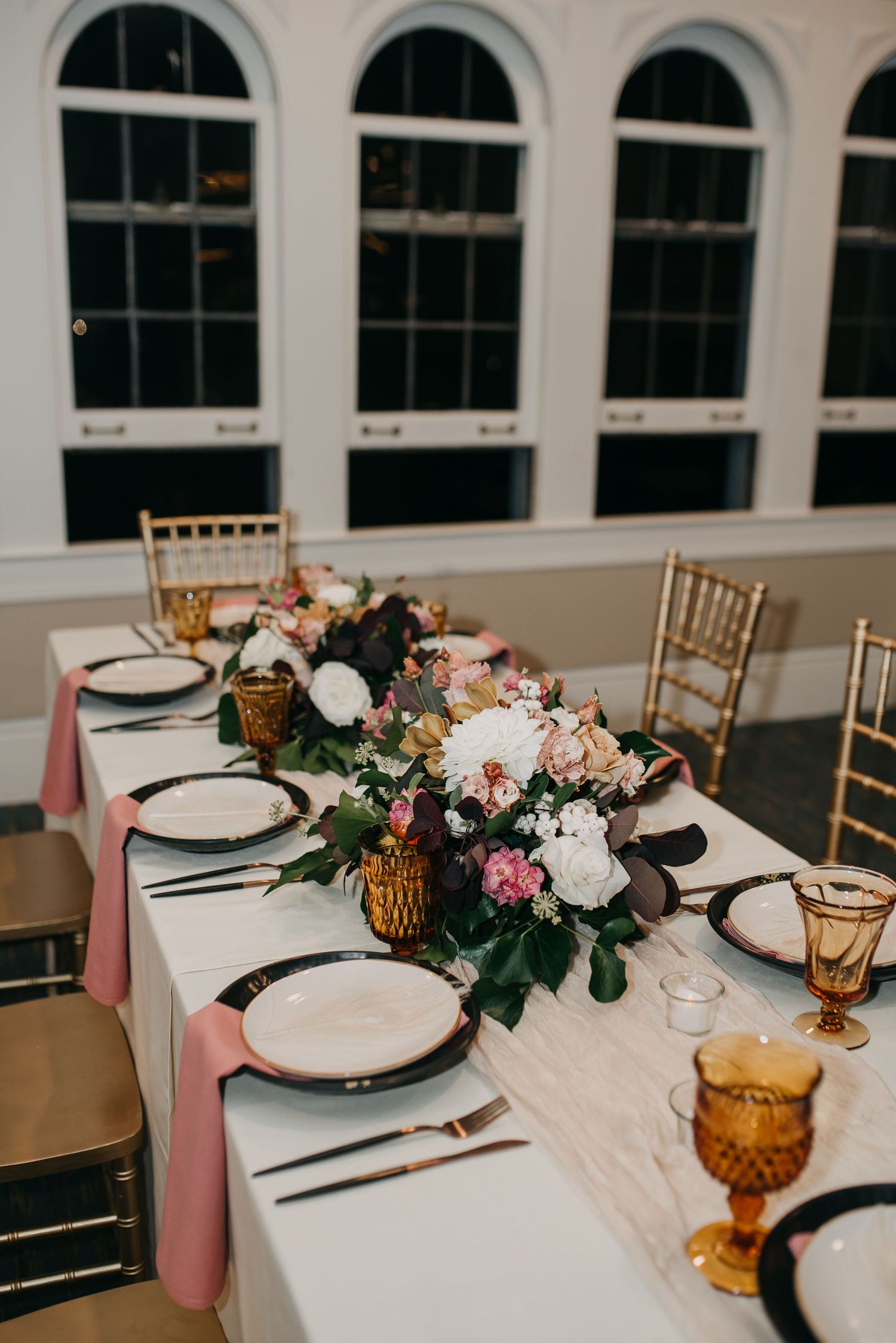 Wedding reception table with pink napkins, amber goblets, gold chiavari chairs, and floral runner.