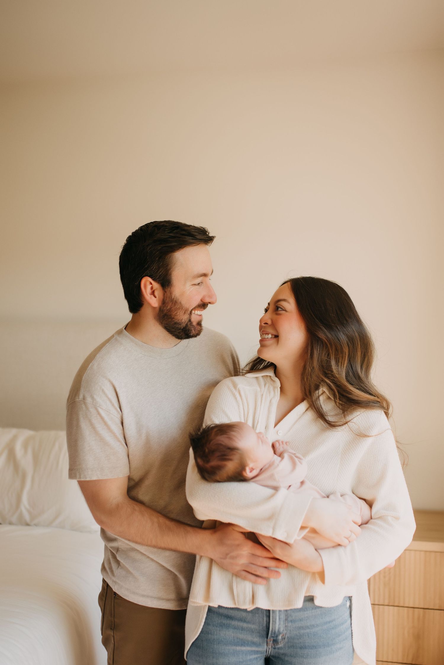 Smiling couple sharing tender moment while holding sleeping newborn baby in bright bedroom.