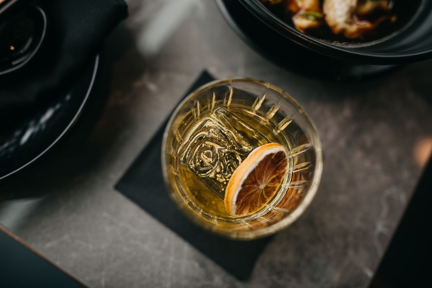 Overhead view of whiskey cocktail with dried orange slice on dark slate coaster.