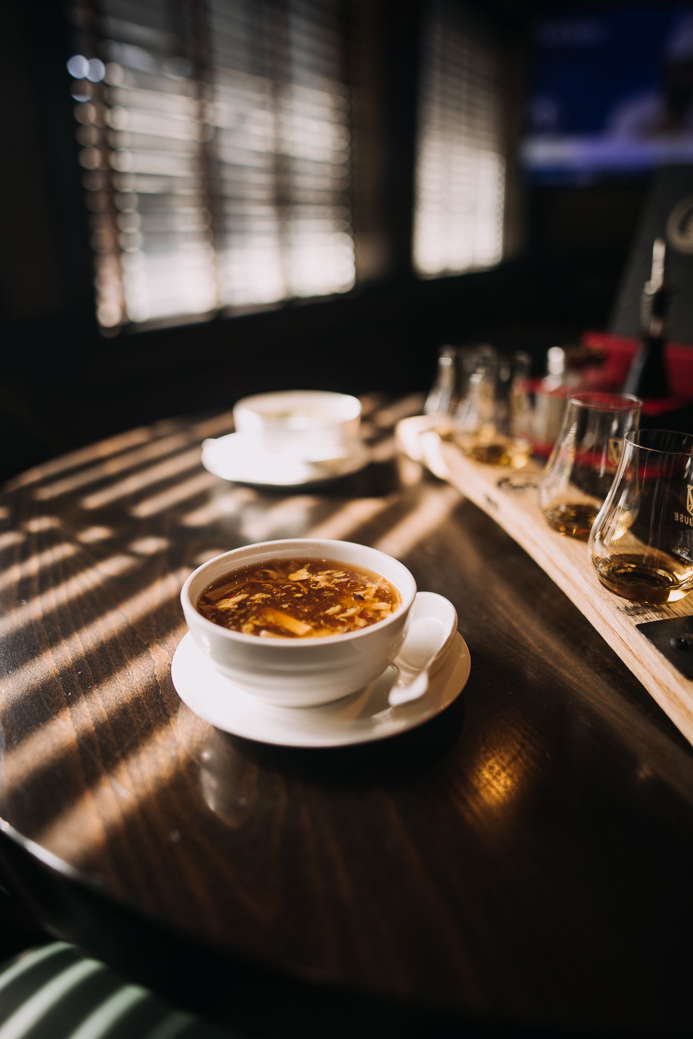 Bowl of soup on saucer beside whiskey flight glasses on sunlit wooden table.