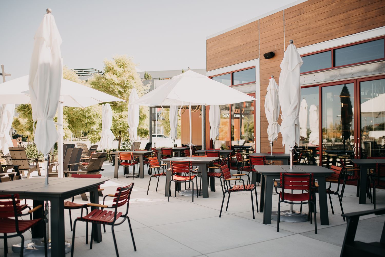 Sunny restaurant patio with red chairs, dark tables and white umbrellas outside modern building.