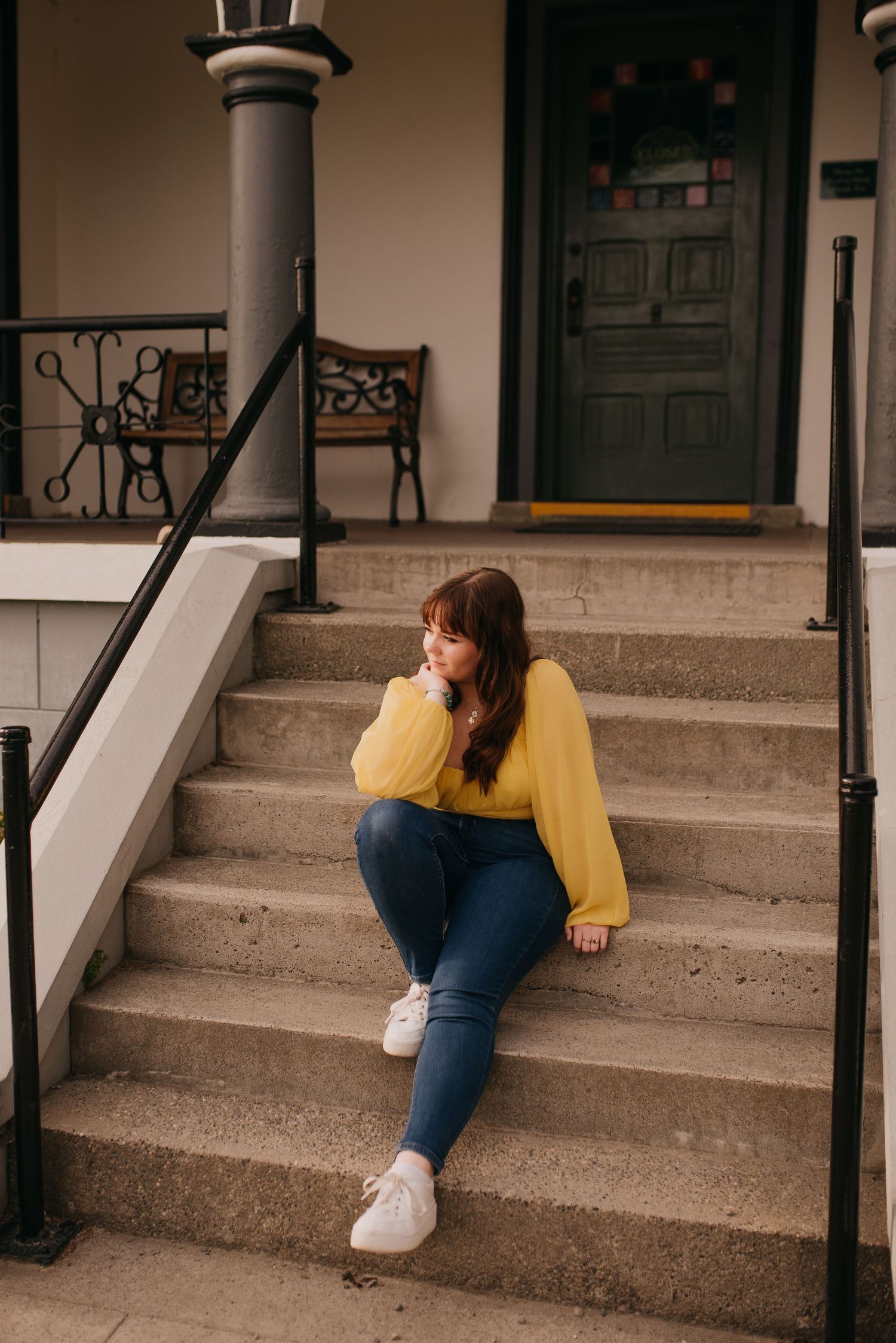 Young woman in yellow blouse and jeans sits thoughtfully on concrete steps outside a historic building.