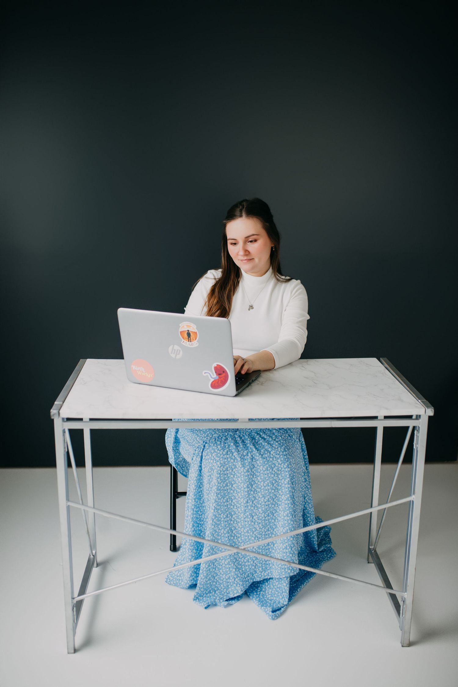 Young woman in white sweater and blue floral skirt working on a sticker-covered laptop at a marble desk.