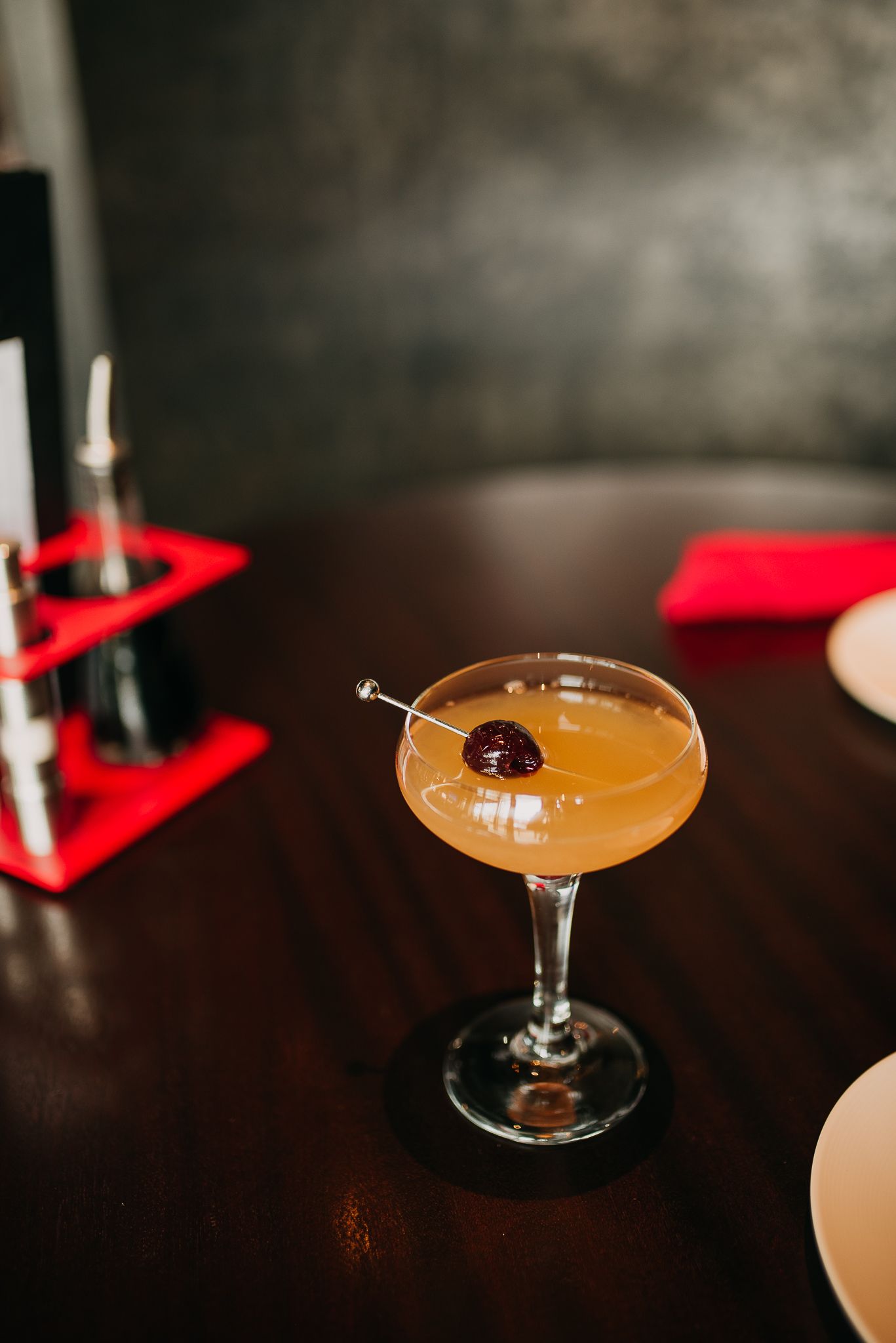 Golden cocktail in a coupe glass garnished with a cherry on a skewer on a dark wood bar.