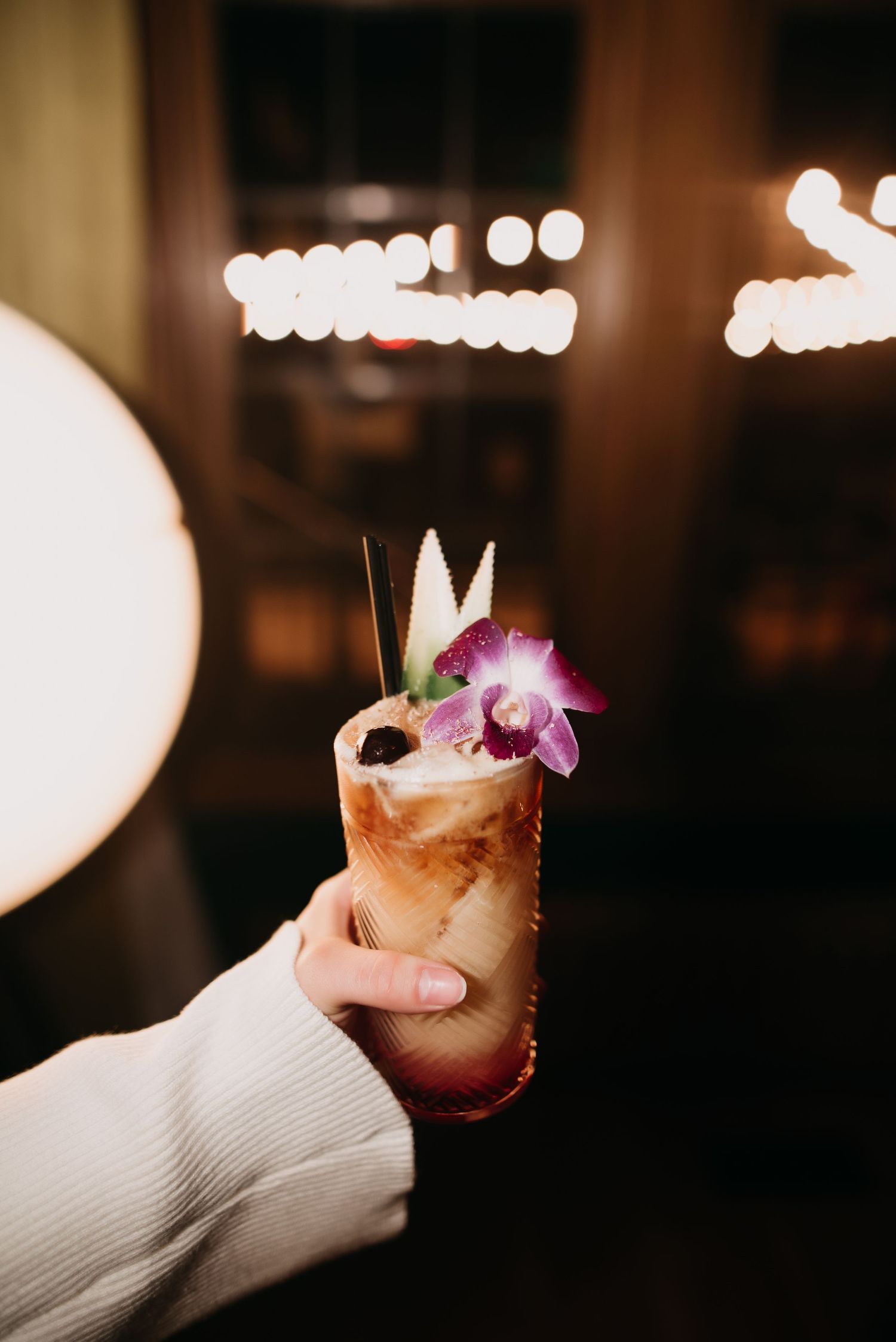 Hand holding tropical cocktail garnished with orchid flower and pineapple leaves in warm bar light.