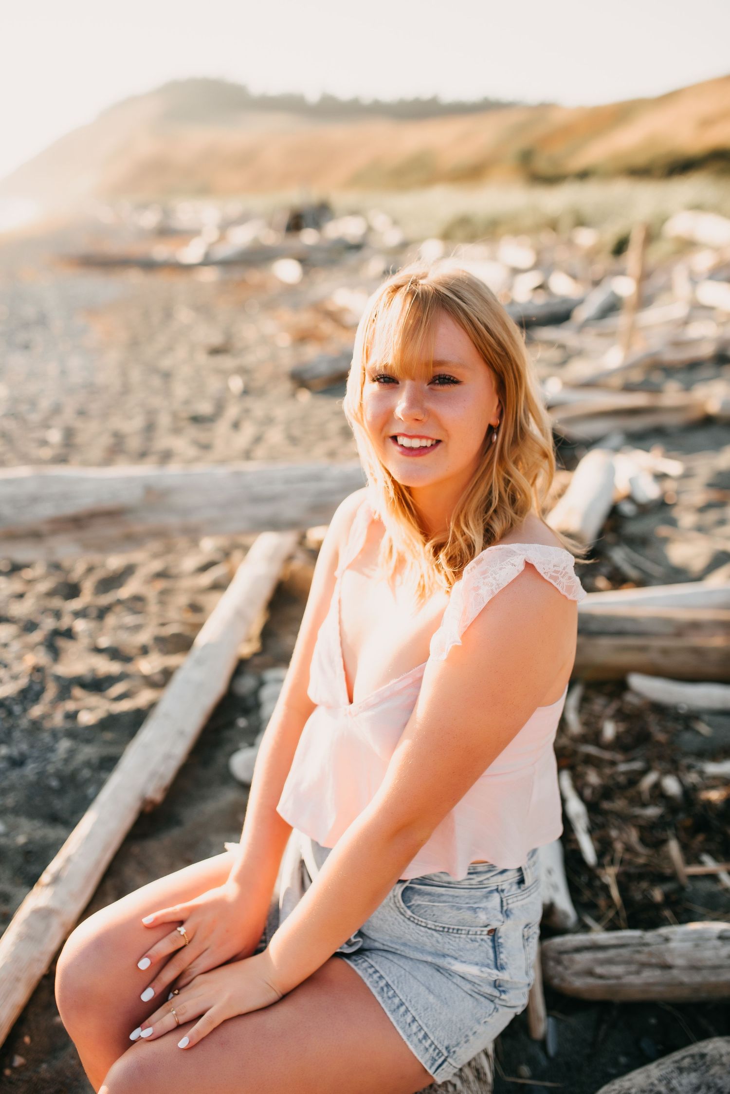 Smiling blonde teen sits on driftwood logs at a sunny beach for senior portrait session.