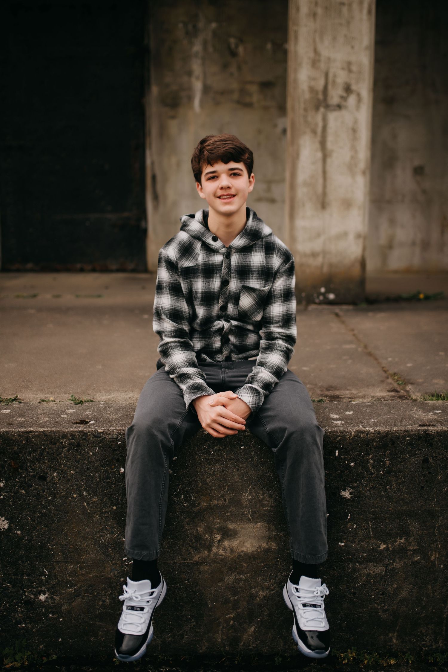 Smiling teenage boy in plaid hoodie sitting on concrete ledge wearing Jordan sneakers.