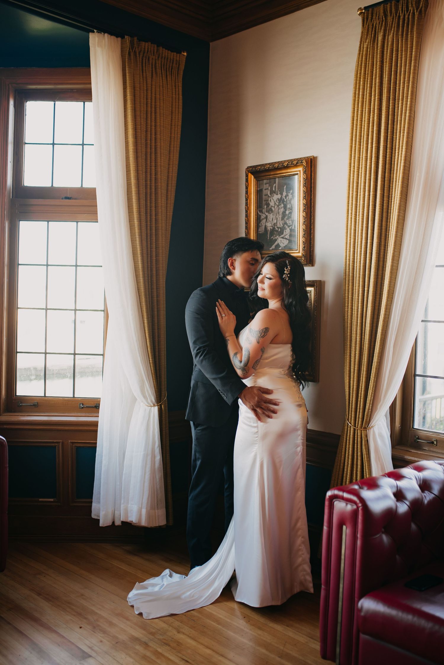 Romantic couple embracing near tall windows in a warmly decorated room with golden drapes and hardwood floors.