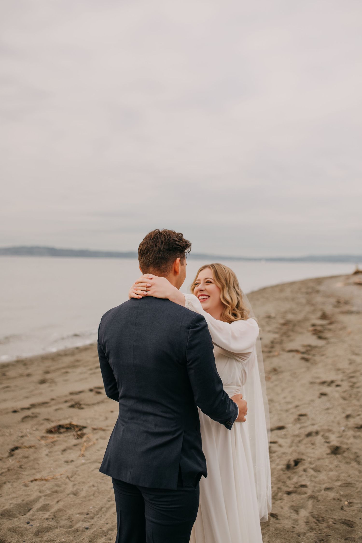 Bride in white gown embracing groom in navy suit on a sandy beach with ocean backdrop.