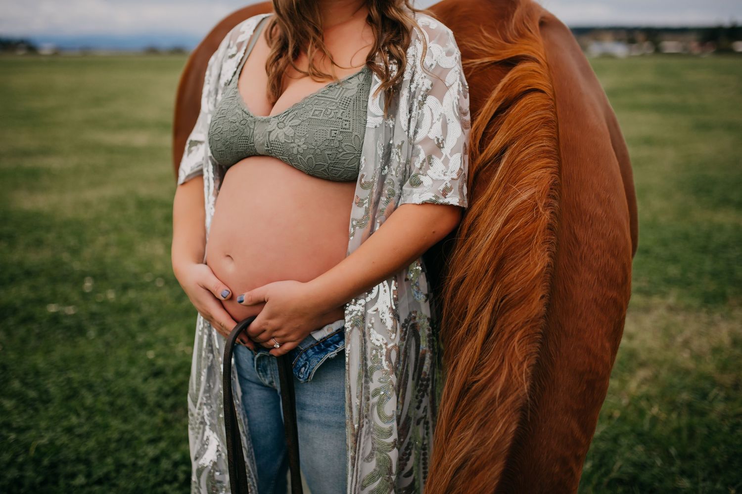 Pregnant woman in sage bralette and jeans standing beside chestnut horse in green pasture.