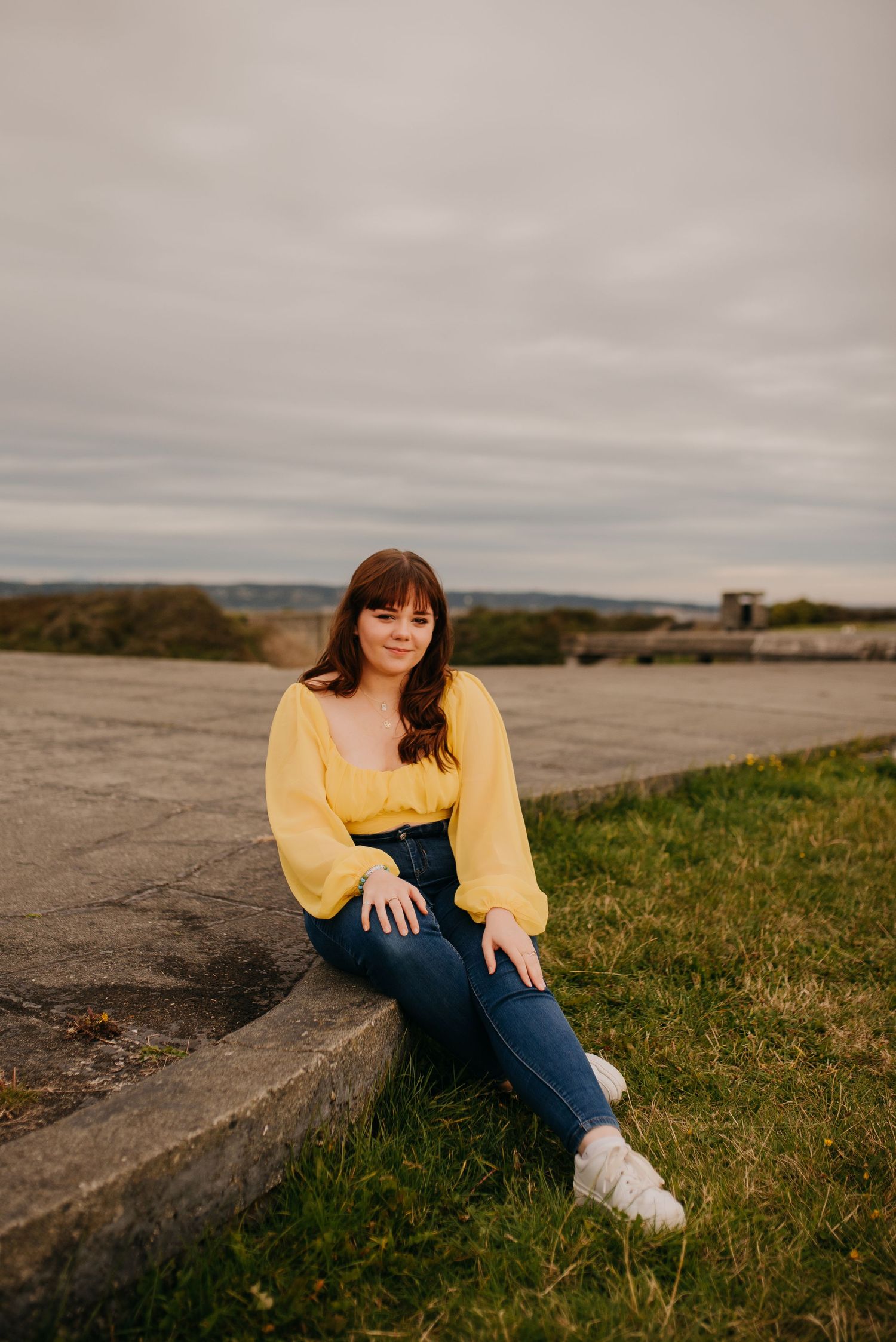 Young woman in yellow blouse and jeans sits on concrete ledge in grassy coastal area under cloudy sky.