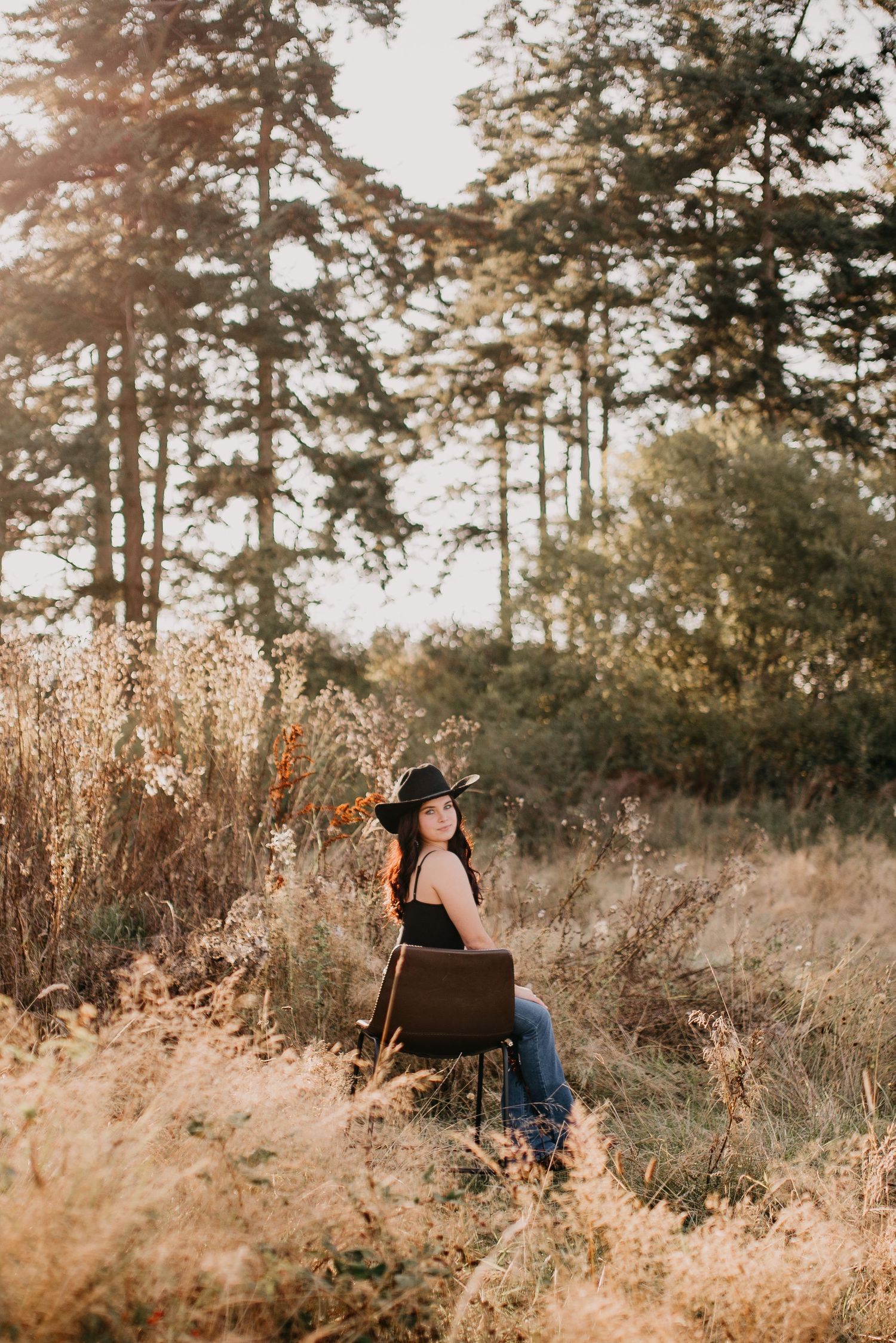 Young woman in black hat sitting on chair in golden field with pine trees at sunset.