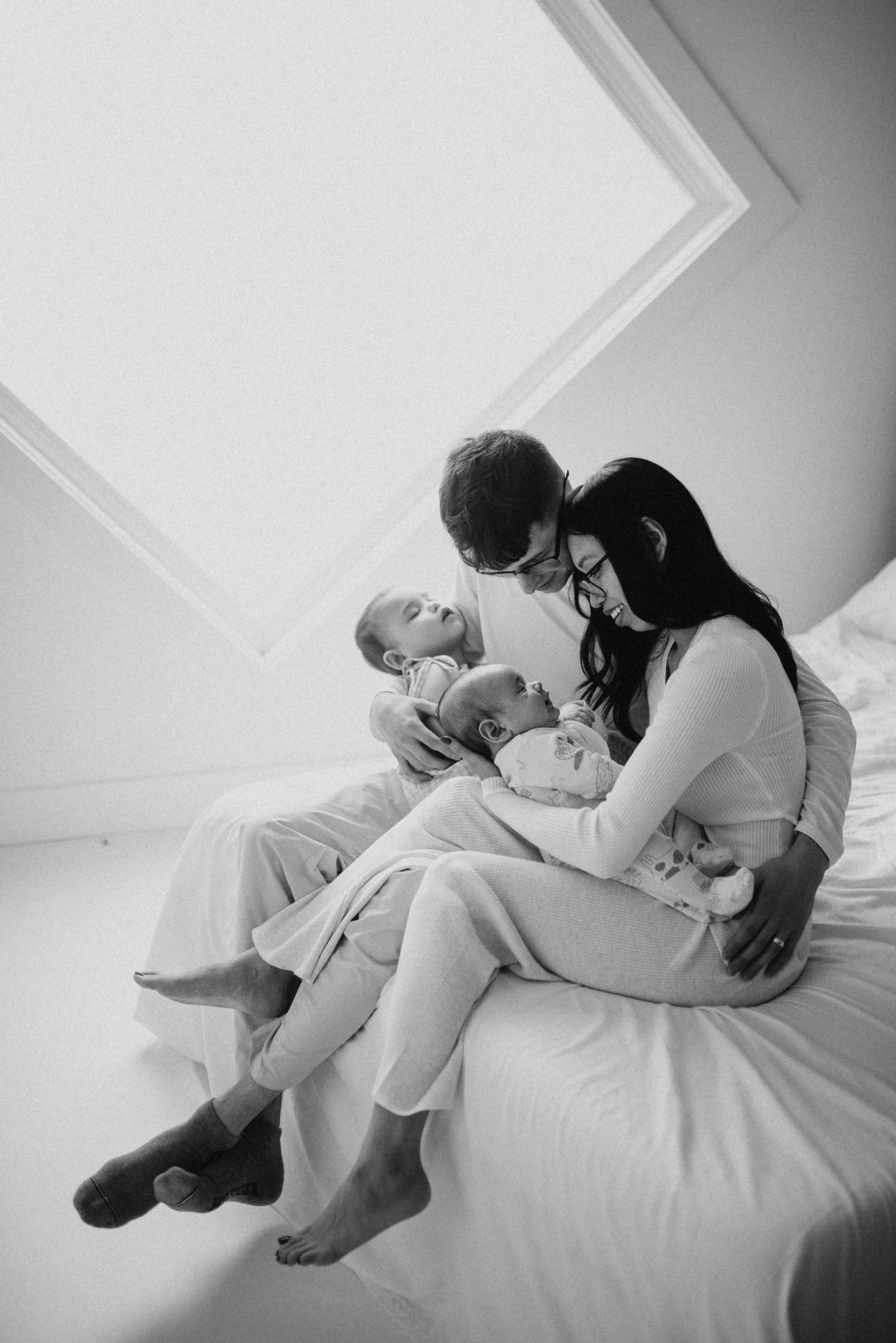 Black and white photo of parents sitting on a bed with twin newborn babies.