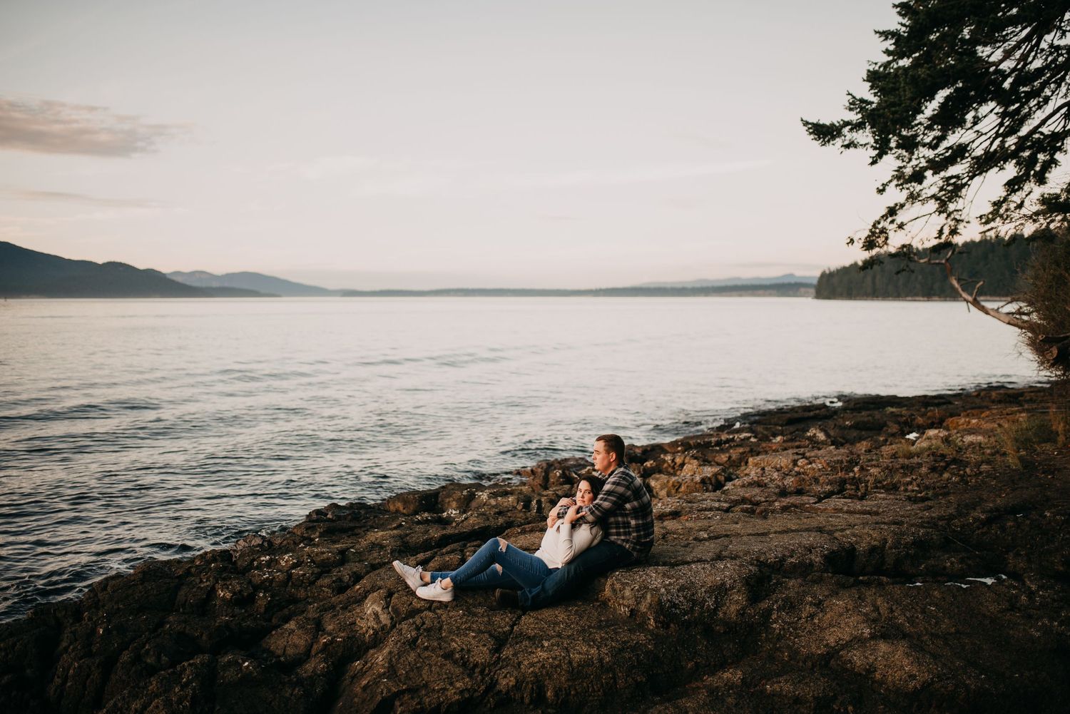 Couple sitting together on rocky shoreline at sunset with calm ocean and mountain views in background.