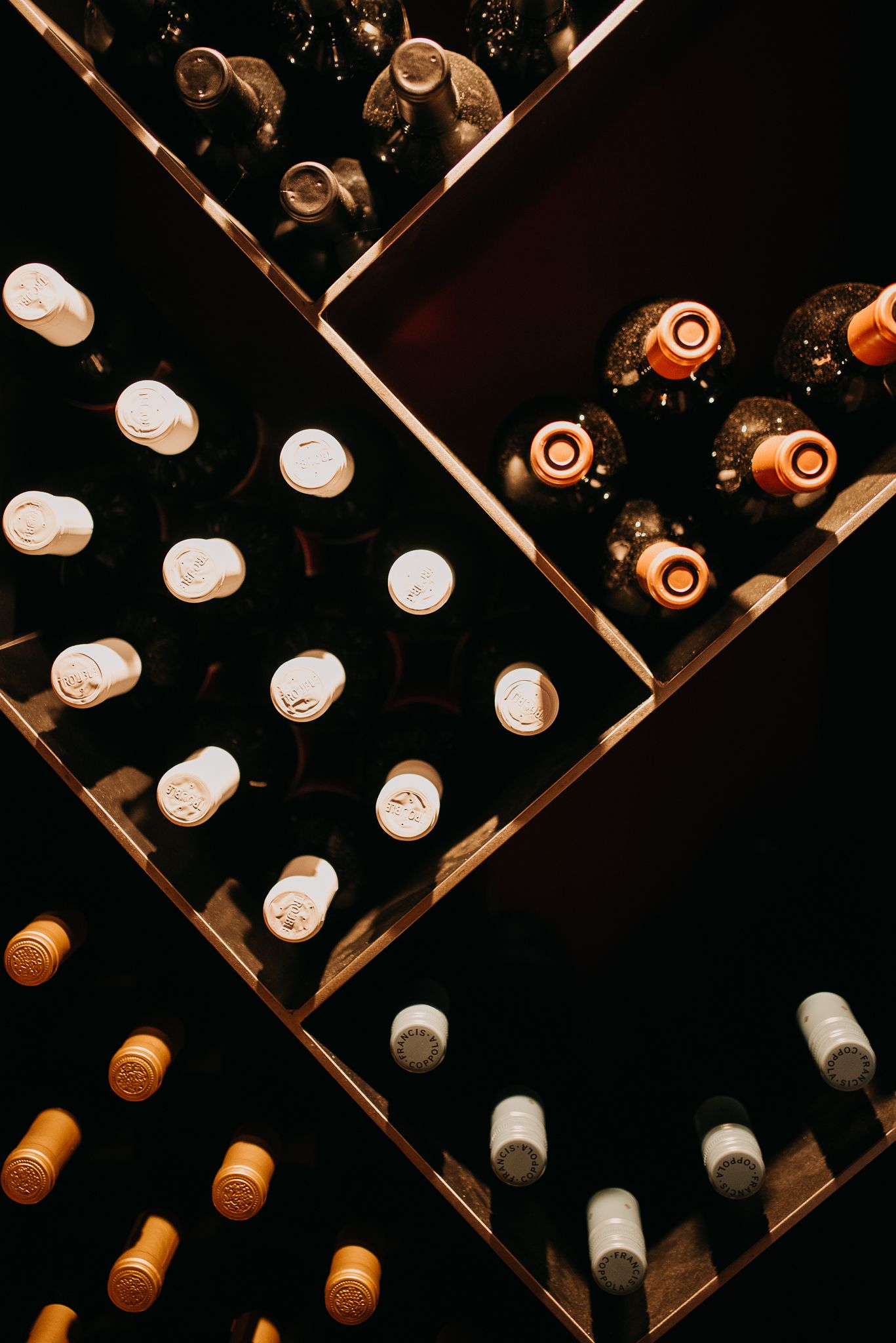 Wine bottles stored horizontally in a dark wooden wine rack, viewed from above showing corks and capsules.