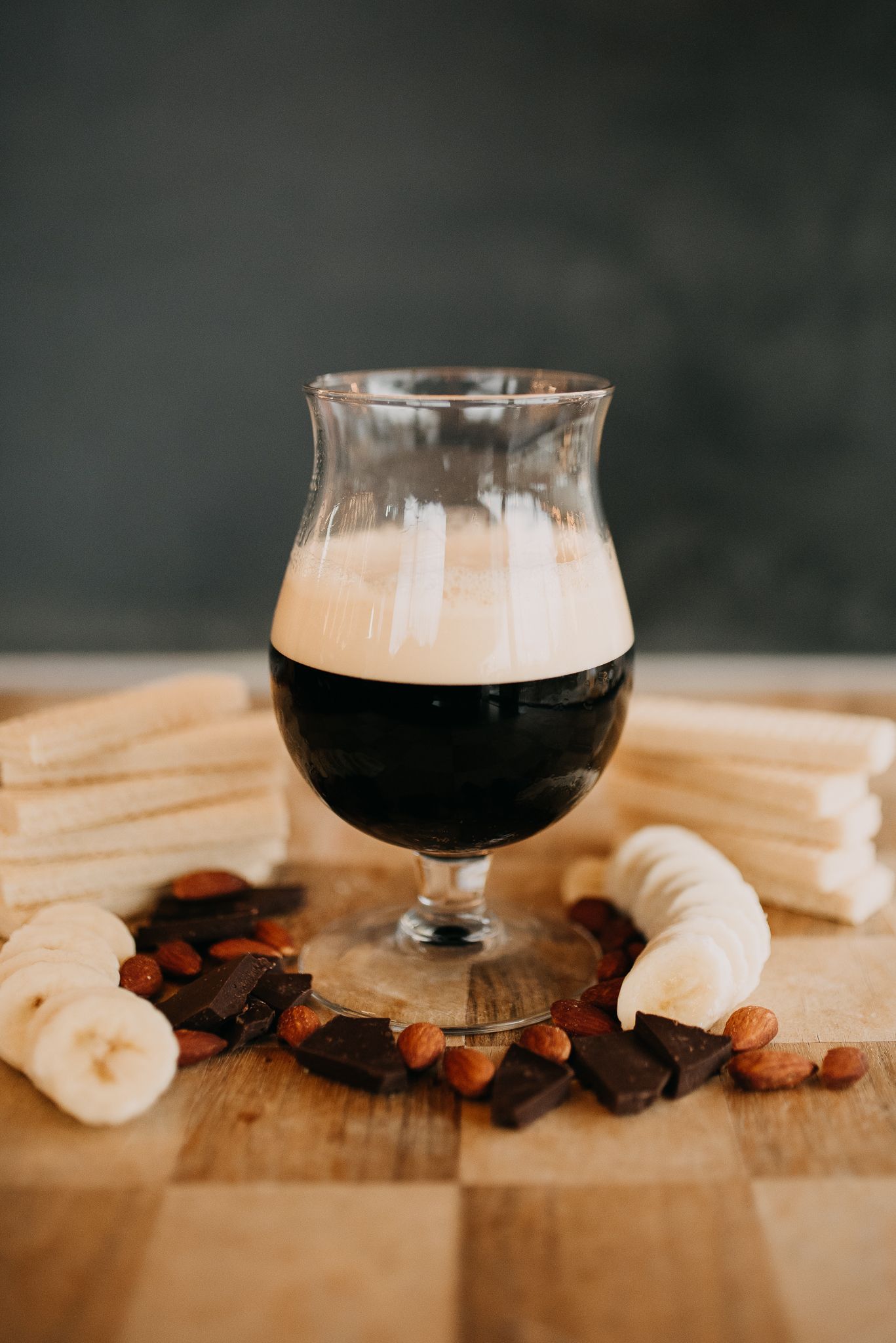 Dark stout beer in a tulip glass surrounded by bananas, almonds, and chocolate pieces.