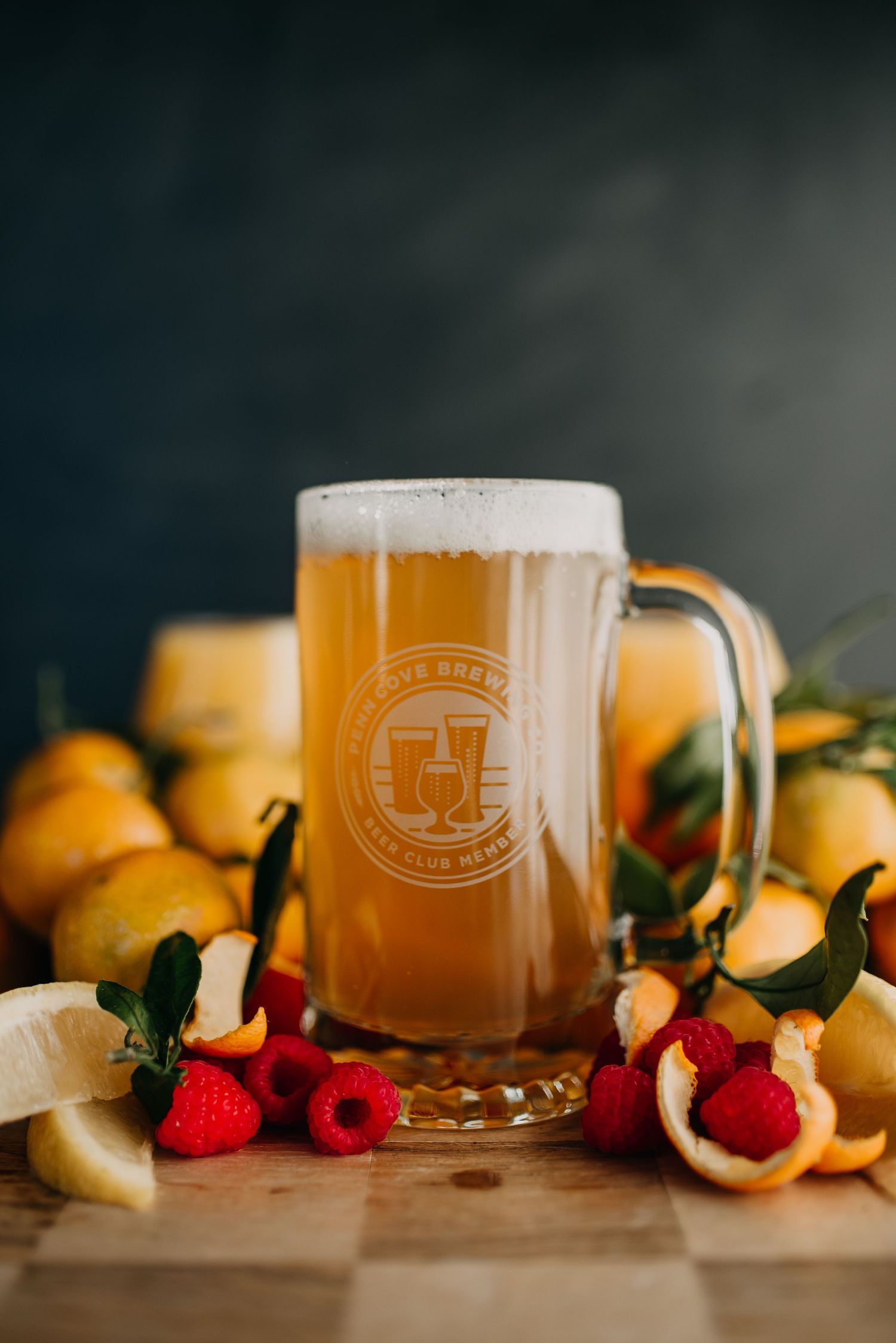 Glass beer mug with frothy ale surrounded by fresh lemons and raspberries on wooden board.