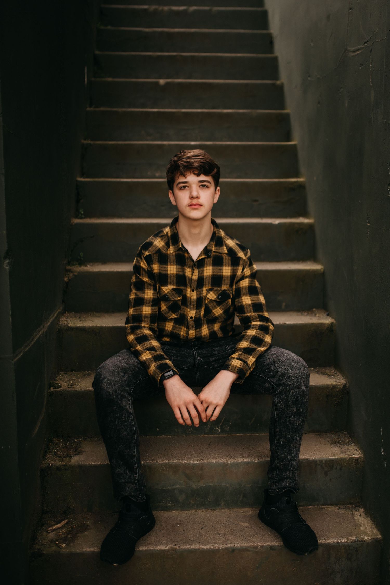 Teenage boy in yellow plaid shirt sitting on concrete stairs in a dimly lit stairwell.