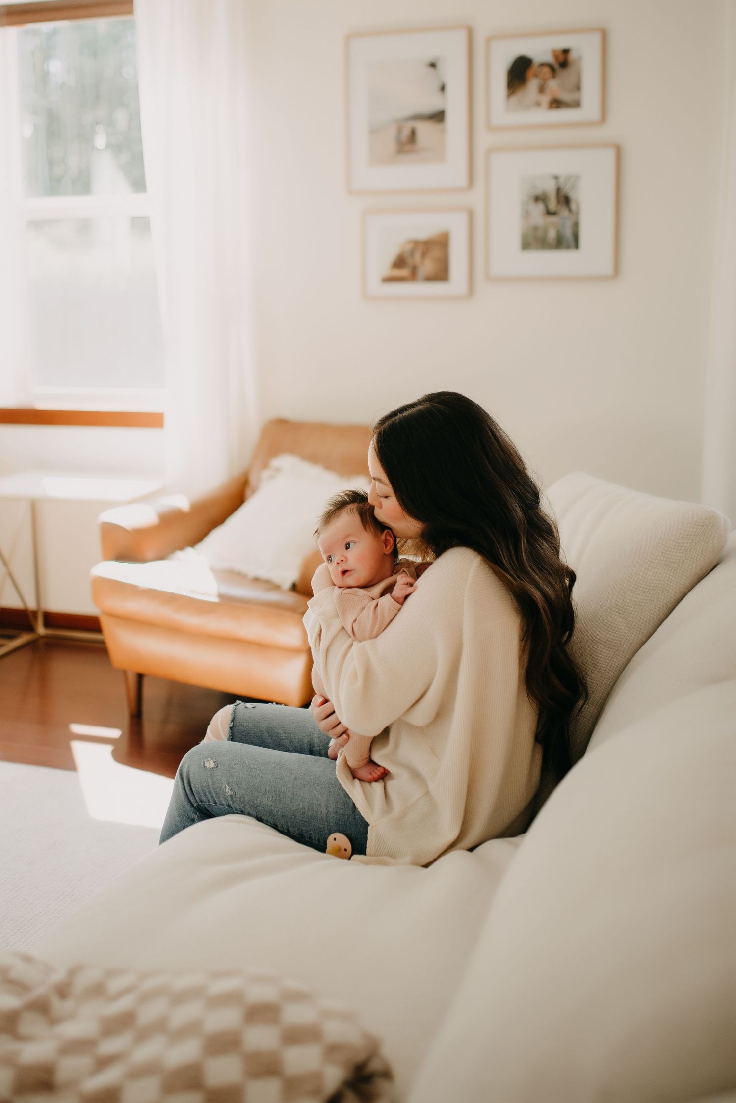Dark-haired mother gently kissing newborn baby while seated on cream sofa in bright living room.