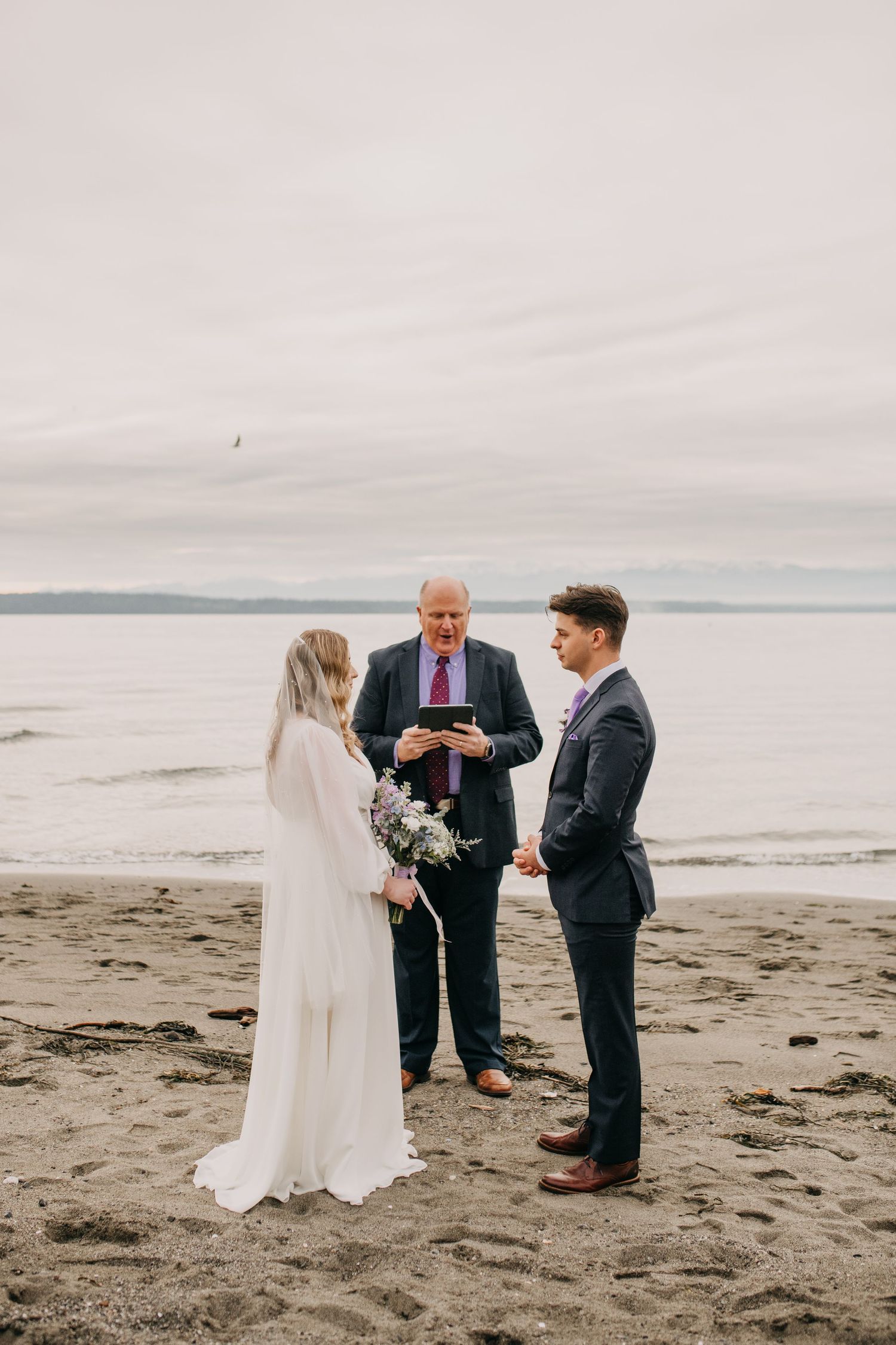 Beach wedding ceremony with bride, groom, and officiant exchanging vows at the ocean shoreline.