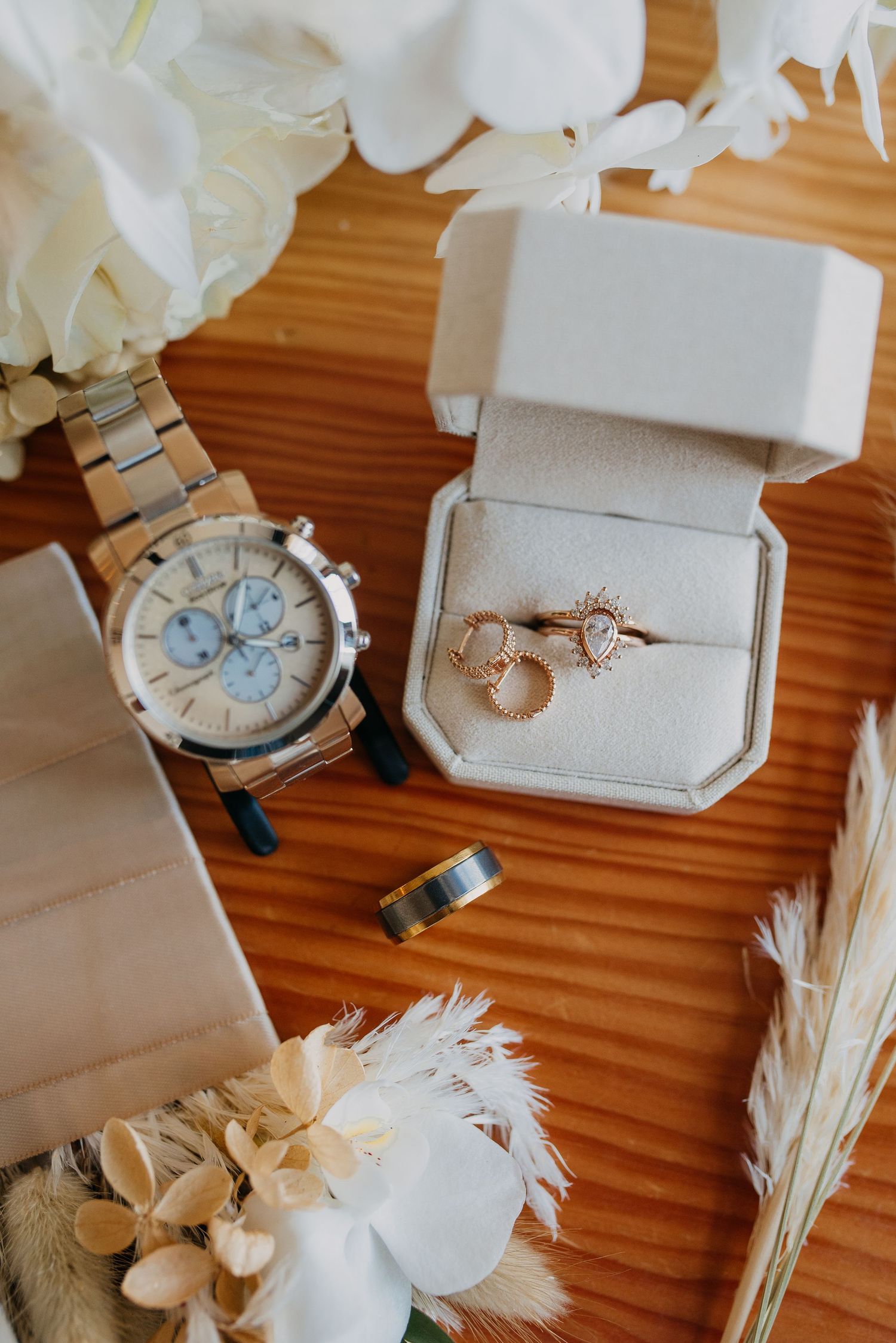 Flatlay of wedding details including rose gold rings in a box, a chronograph watch, and floral accents.