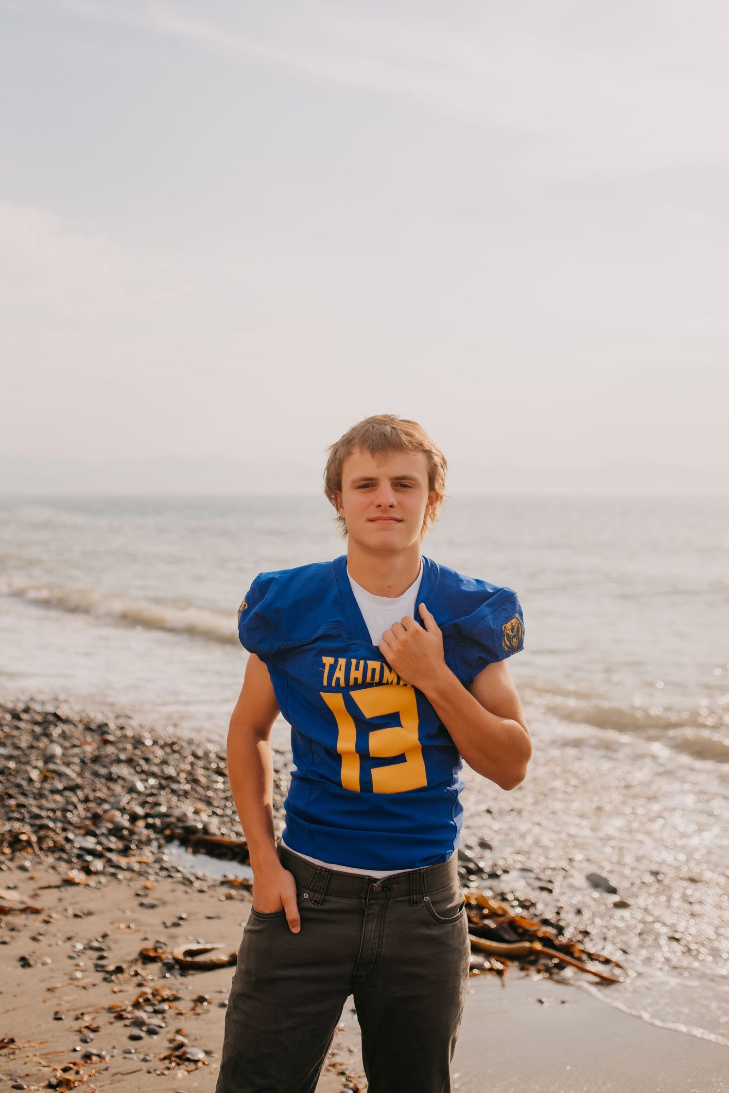 Young male athlete in blue Tahoe football jersey number 13 stands on rocky beach shoreline.