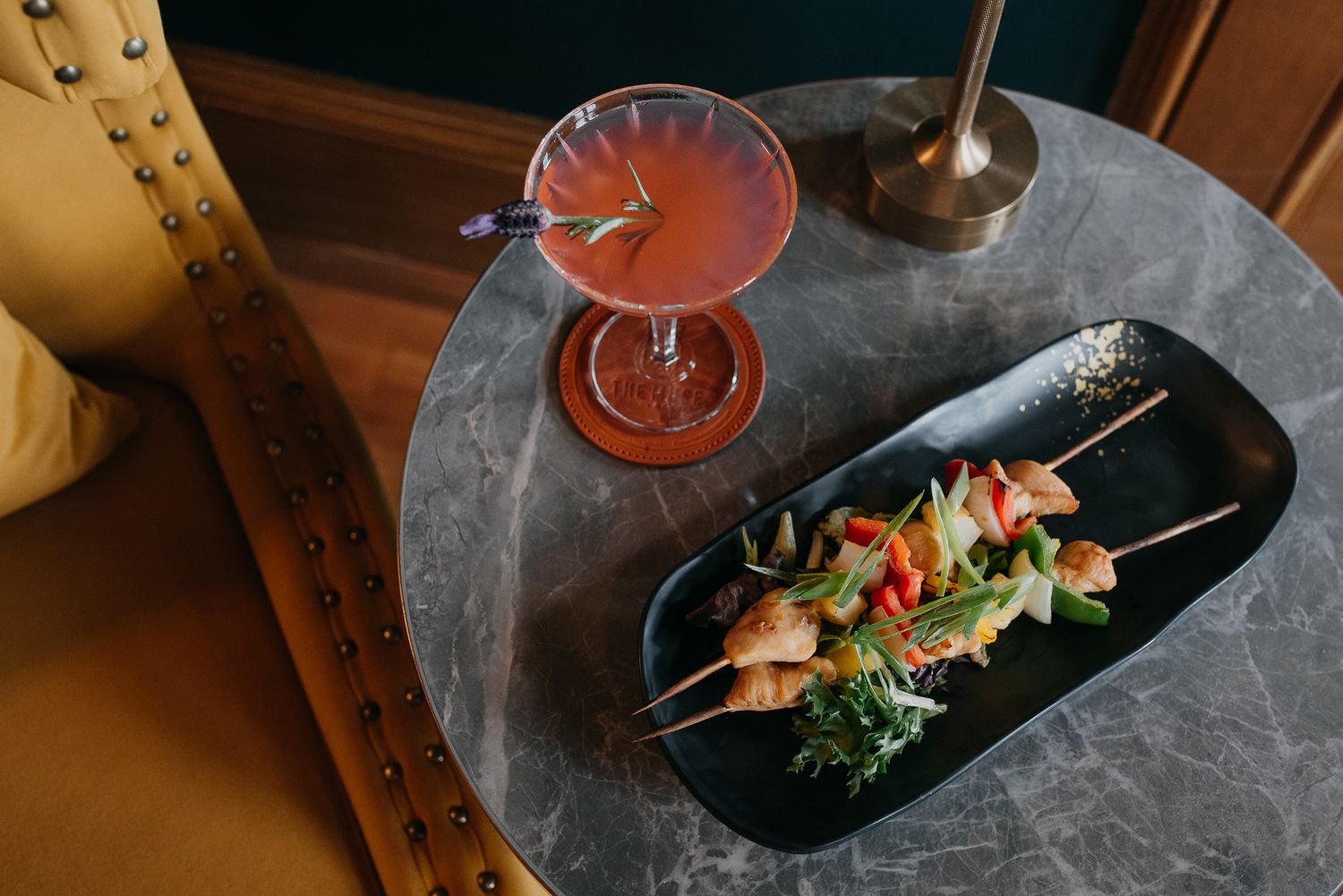 Pink cocktail in coupe glass beside skewers with vegetables on black plate on marble table.