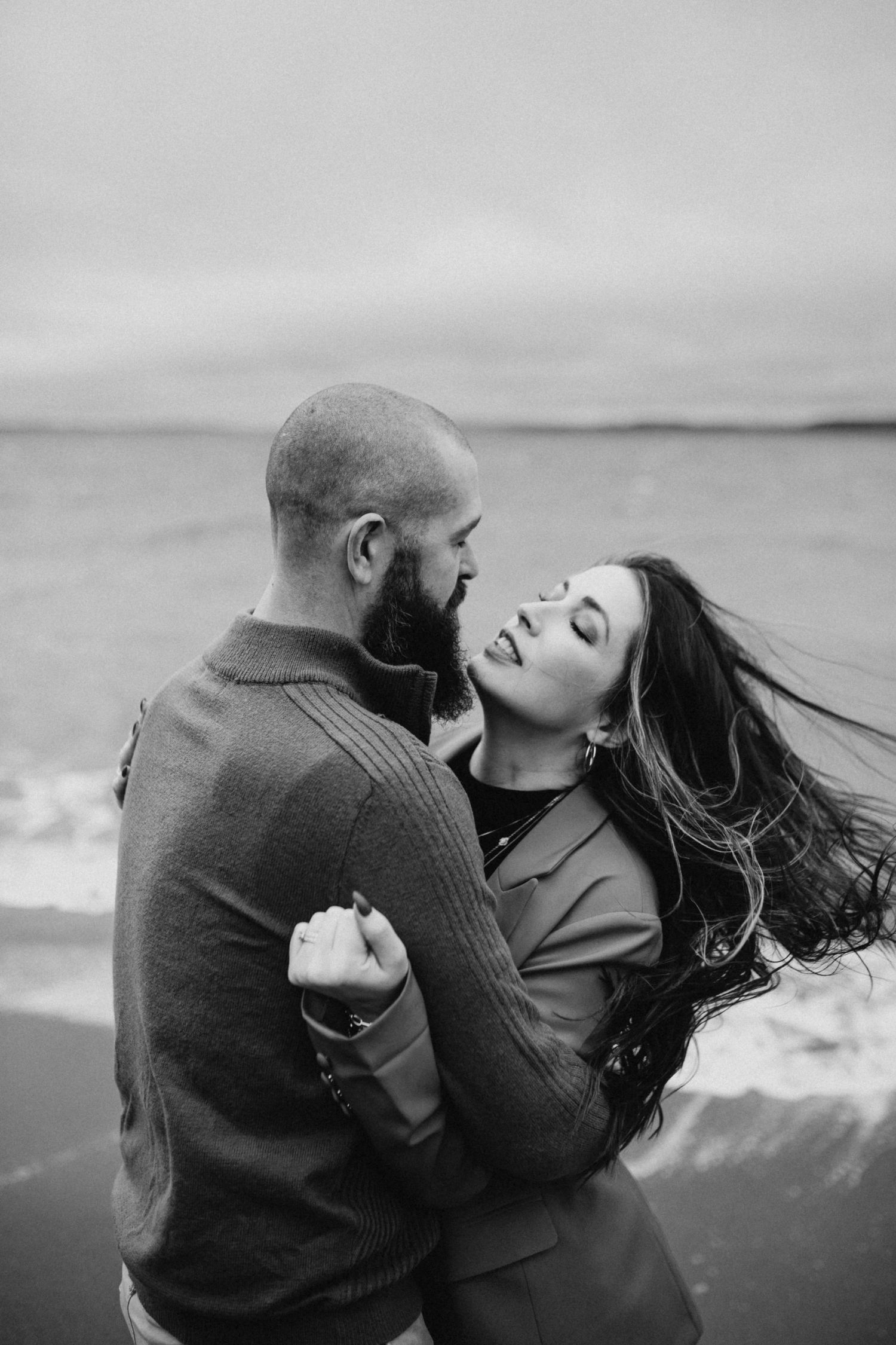 Romantic couple embracing on windy beach shoreline, black and white coastal engagement photography.