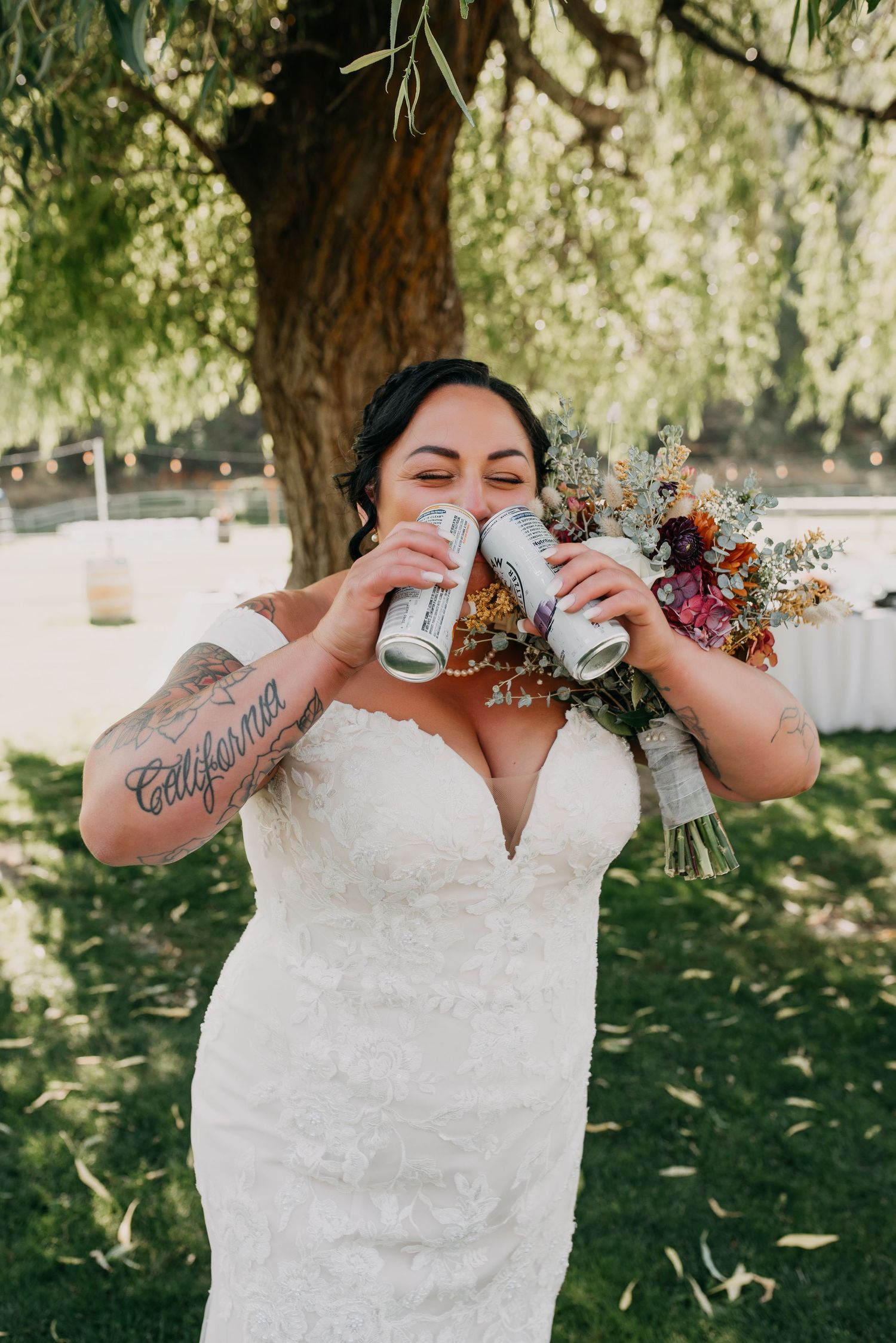 Tattooed bride in lace gown holds bouquet and beer cans under a willow tree at outdoor wedding.