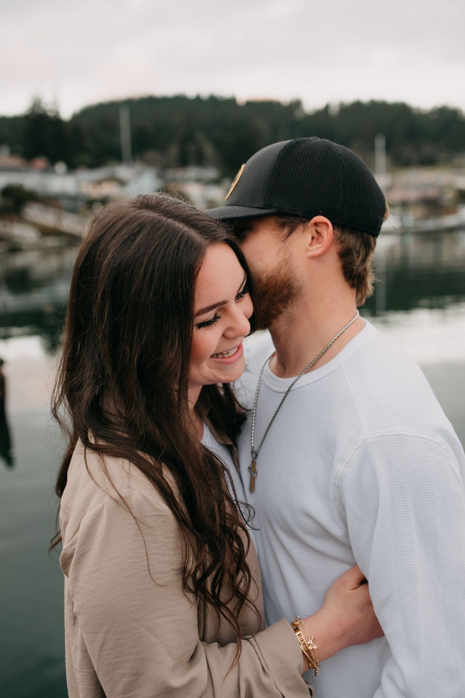 Happy couple sharing a tender moment dockside with marina and trees in background.