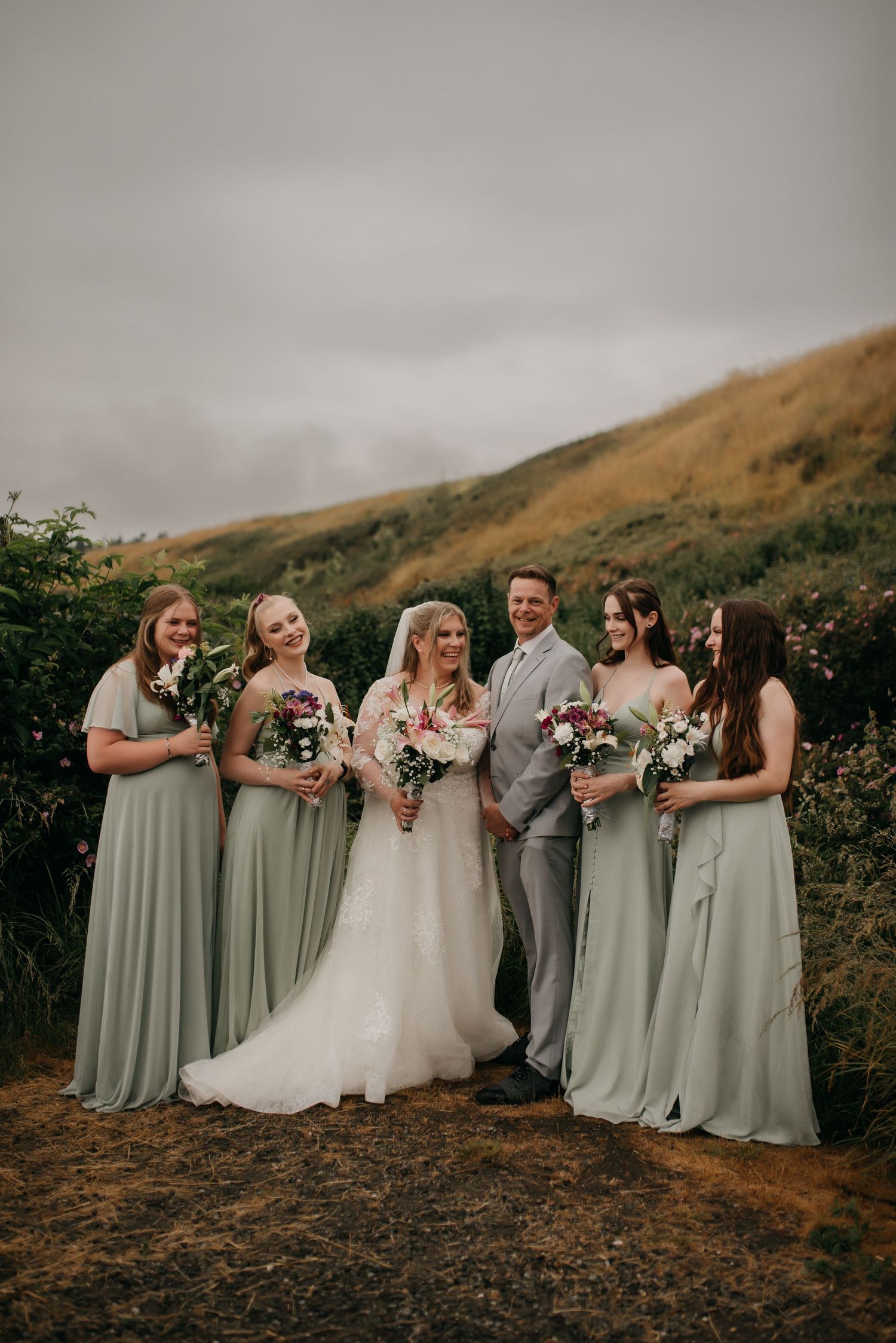 Wedding party poses outdoors with bride in white gown and bridesmaids in sage green dresses against rolling hills.
