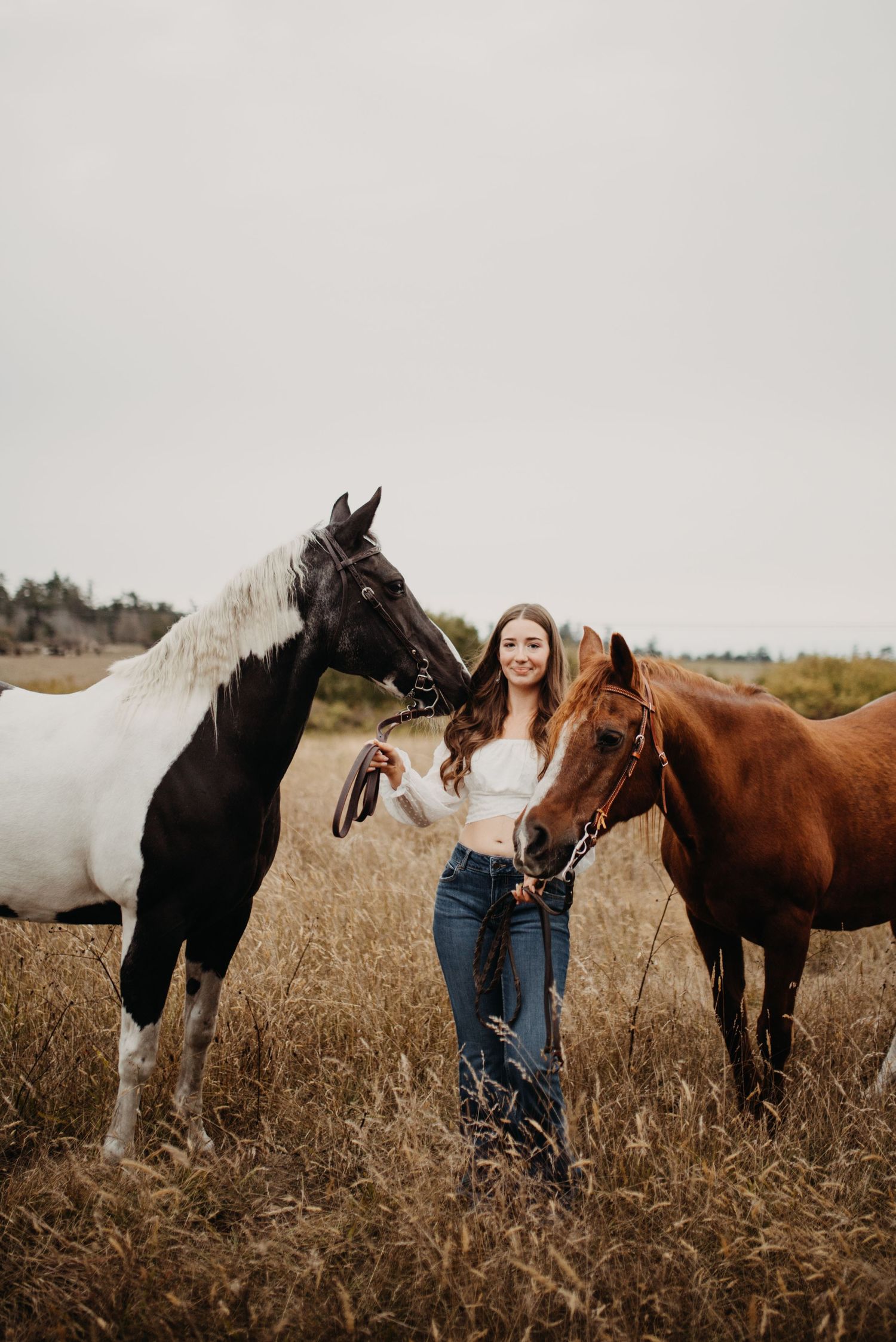 Young woman in white crop top and jeans standing between two horses in autumn field.