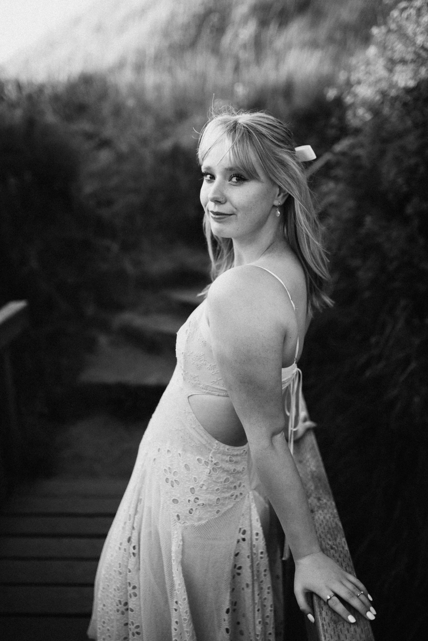 Young woman in floral dress poses on wooden walkway surrounded by lush greenery in black and white photo.