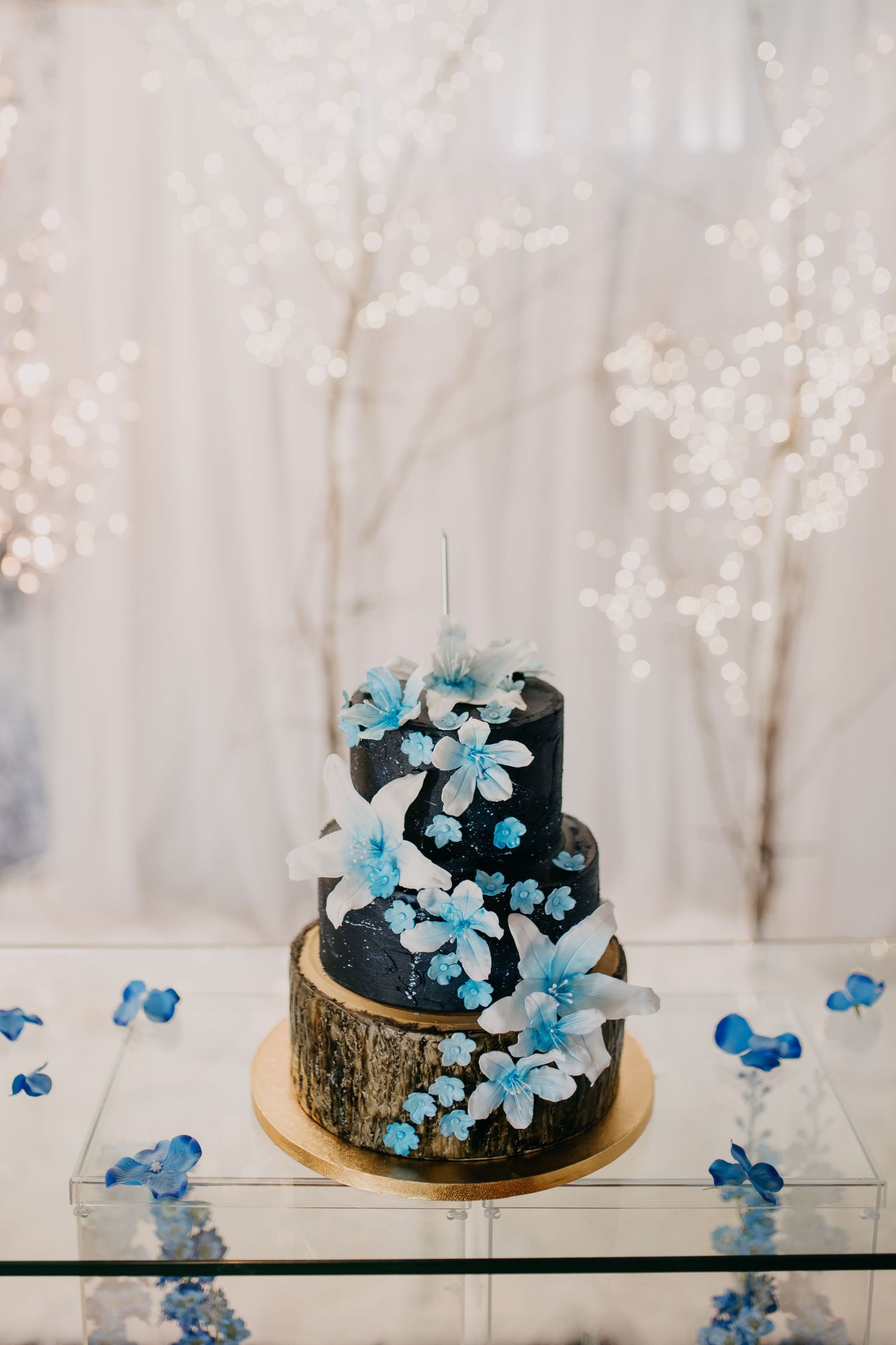 Elegant three-tier black wedding cake decorated with blue and white sugar flowers on a glass table.