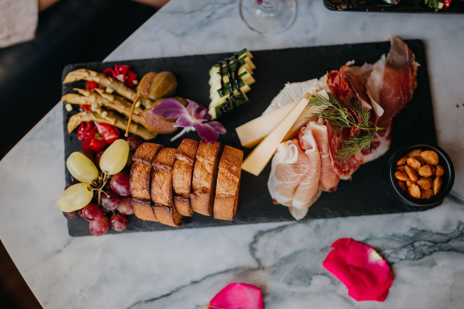 Elegant charcuterie board with meats, cheeses, bread, grapes, and garnishes on a black slate platter.