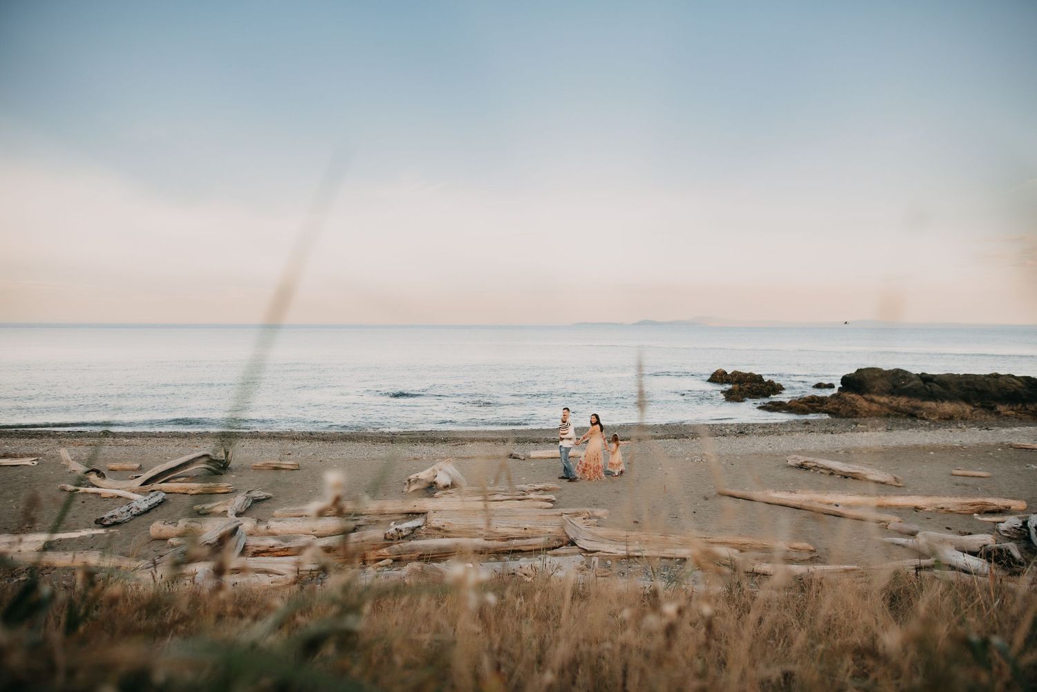 Couple walking hand in hand on driftwood-lined beach at sunset with calm ocean waves.