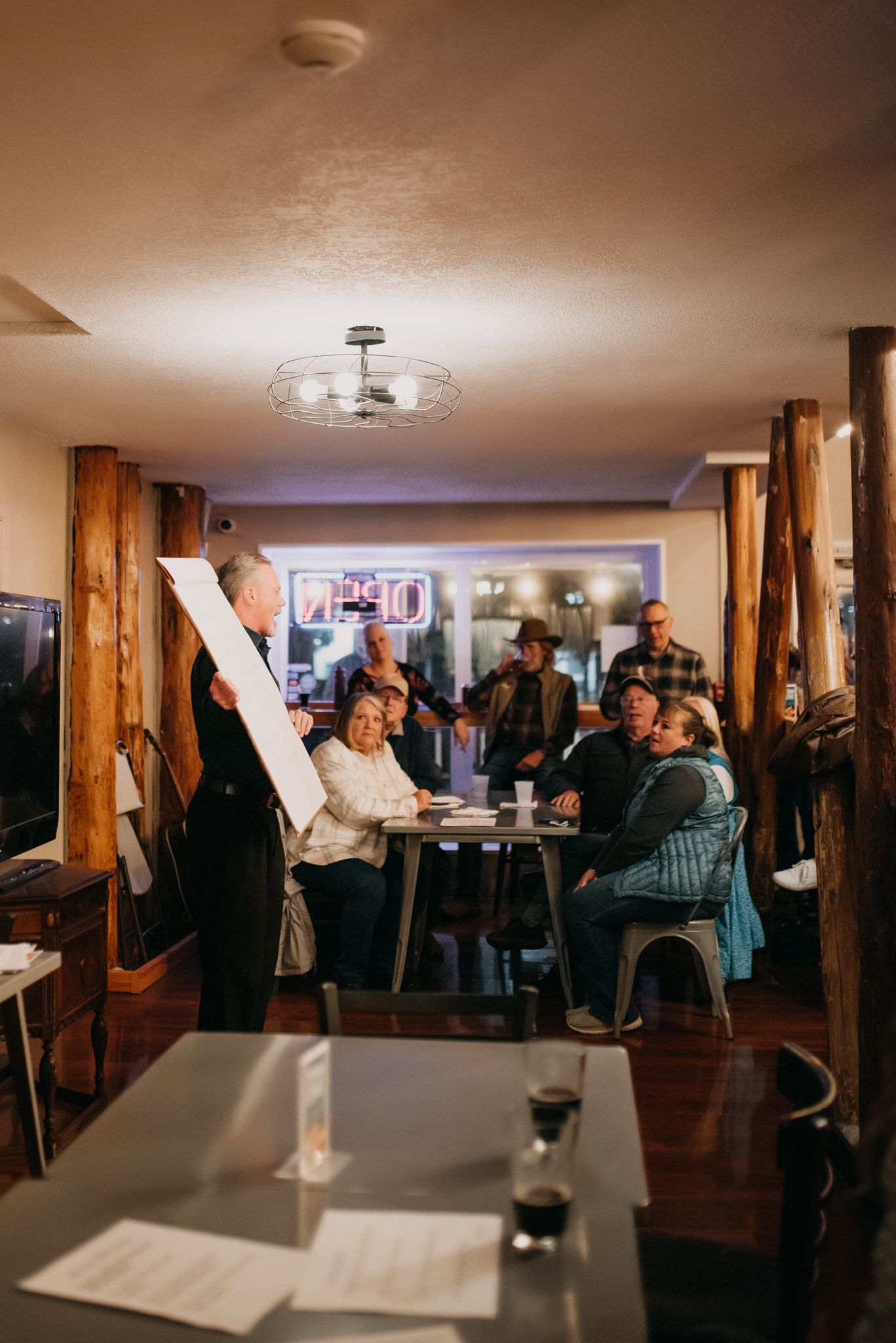Cozy restaurant interior with warm lighting, wooden pillars, and guests enjoying an evening gathering or presentation.