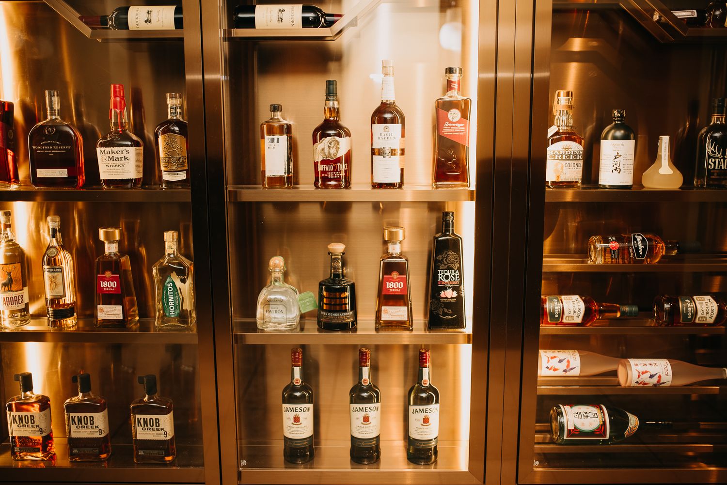 A well-stocked bar shelf displaying an assortment of premium liquor bottles in a warmly lit cabinet.