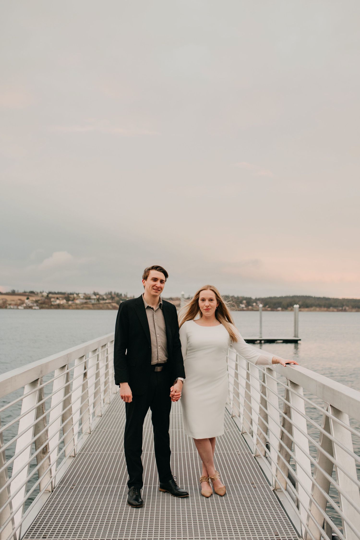 Engaged couple holding hands on a white pier overlooking calm ocean waters at sunset.
