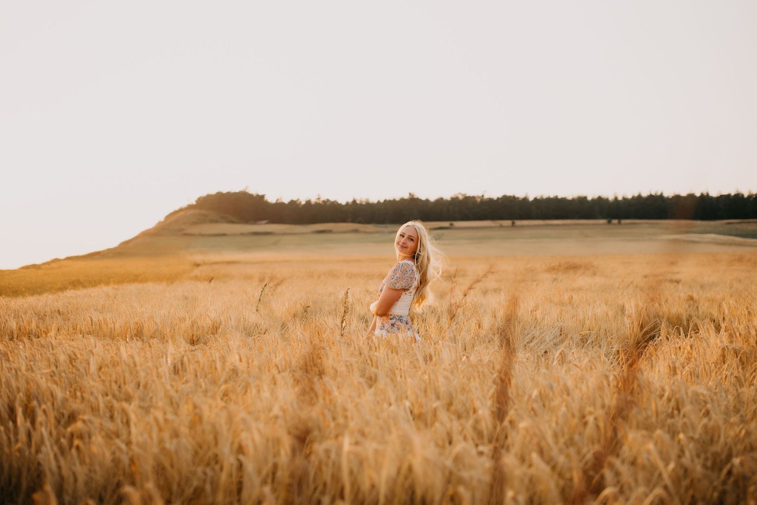 Blonde woman in floral dress walks through a golden wheat field with a tree-lined hill in the background.