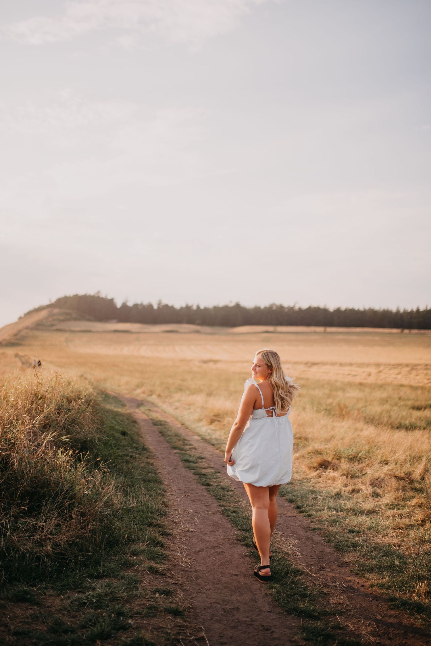 Blonde woman in white sundress walks along rural dirt path in golden sunset light.