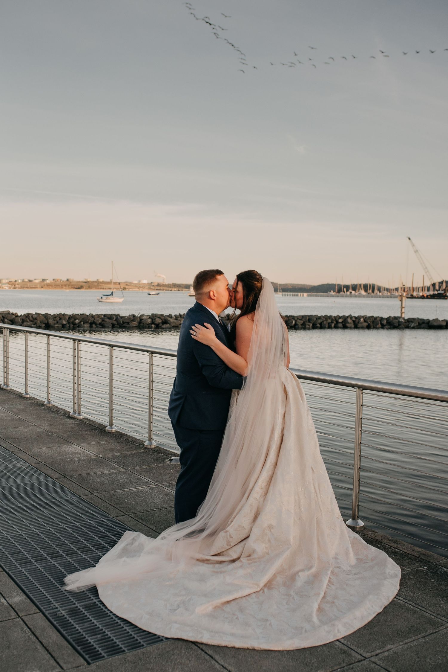 Bride and groom share a kiss on a waterfront pier at golden hour with a flowing cathedral veil.